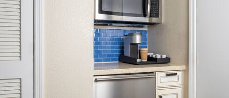 A compact kitchen with a stainless steel microwave, coffee machine, and a blue tiled backsplash.