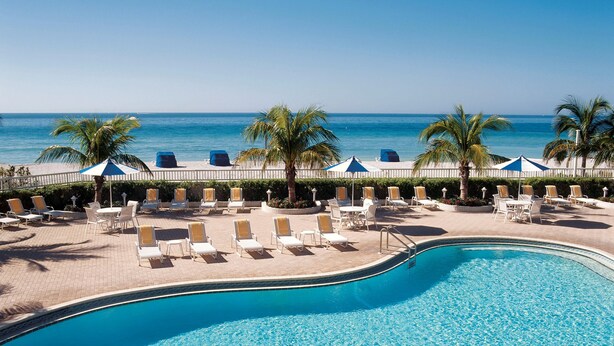 A pool area with lounge chairs and umbrellas, a palm tree, and a view of the ocean. - Lido Beach Resort (Sarasota)