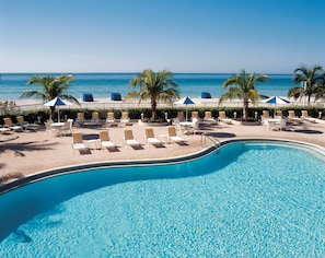 A pool area with lounge chairs and umbrellas, a palm tree, and a view of the ocean. - Lido Beach Resort (Sarasota)