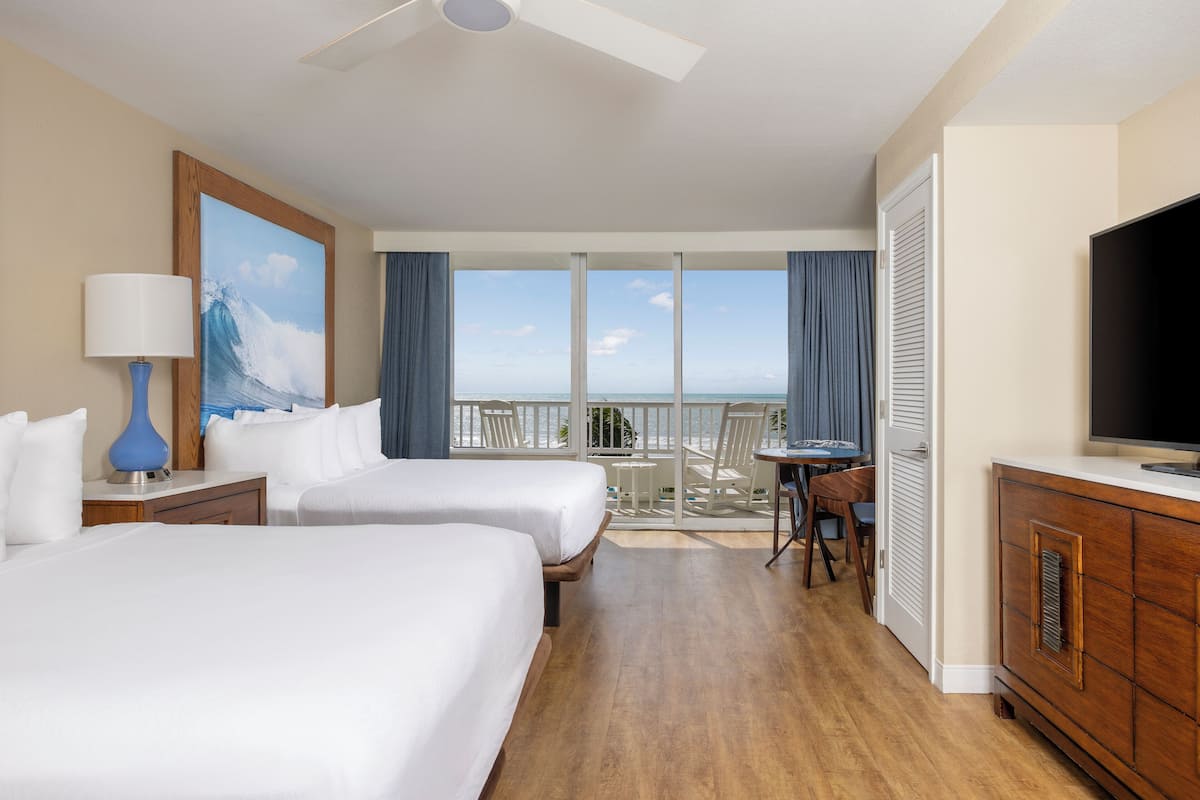 A hotel room with two beds, a TV, a desk, and a balcony with a view of the ocean.