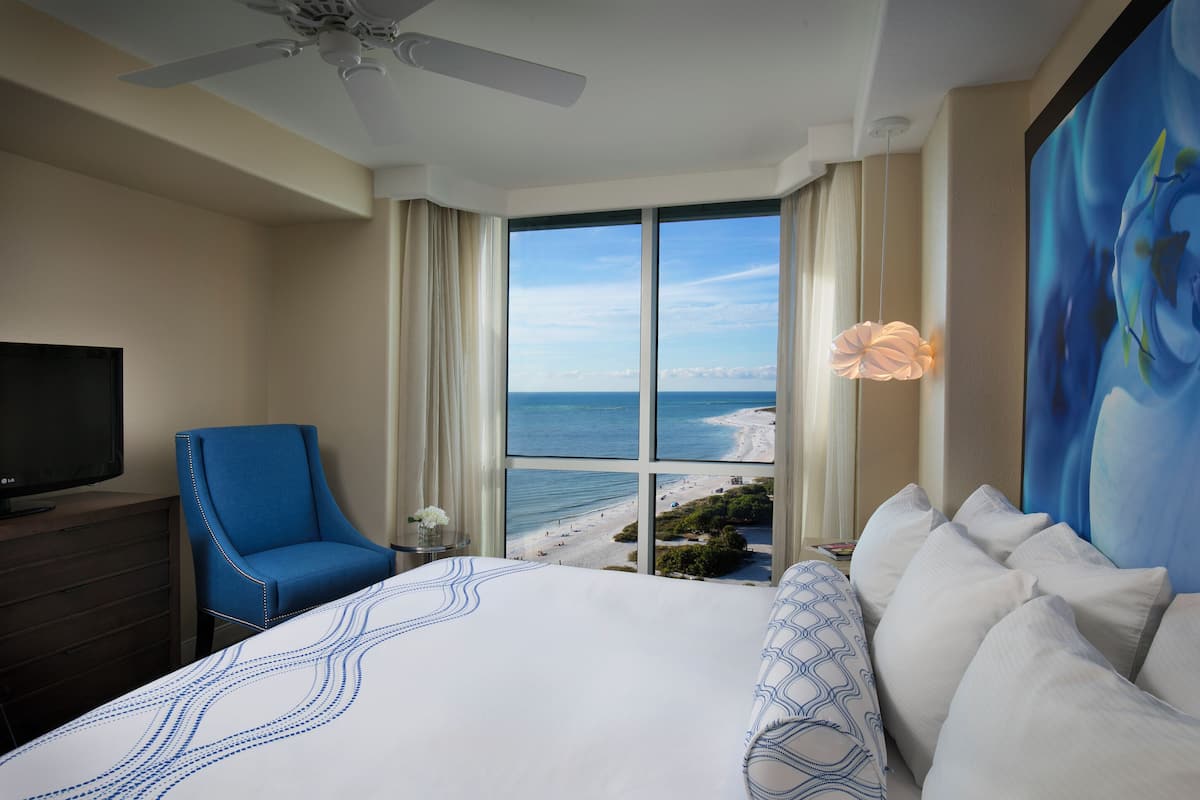 A hotel room with a large bed, a blue chair, a TV, and a view of the beach.