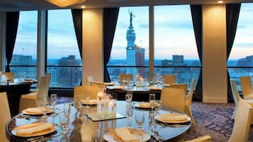 A dining area with tables set for a meal, large windows with a cityscape view, and a statue on a pedestal in the background.