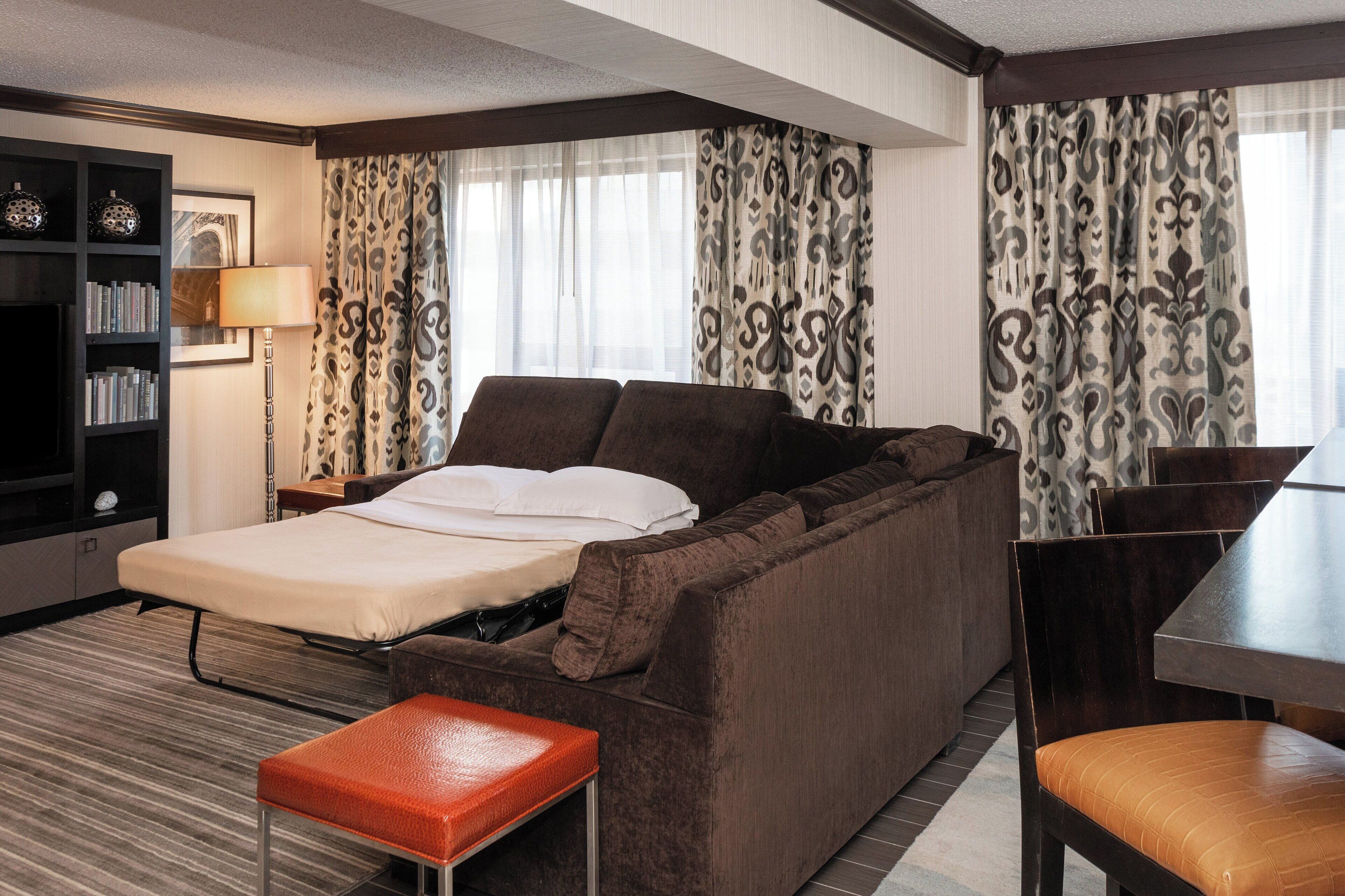 A compact hotel room with a sofa bed, a small table, a bookshelf, and a lamp.