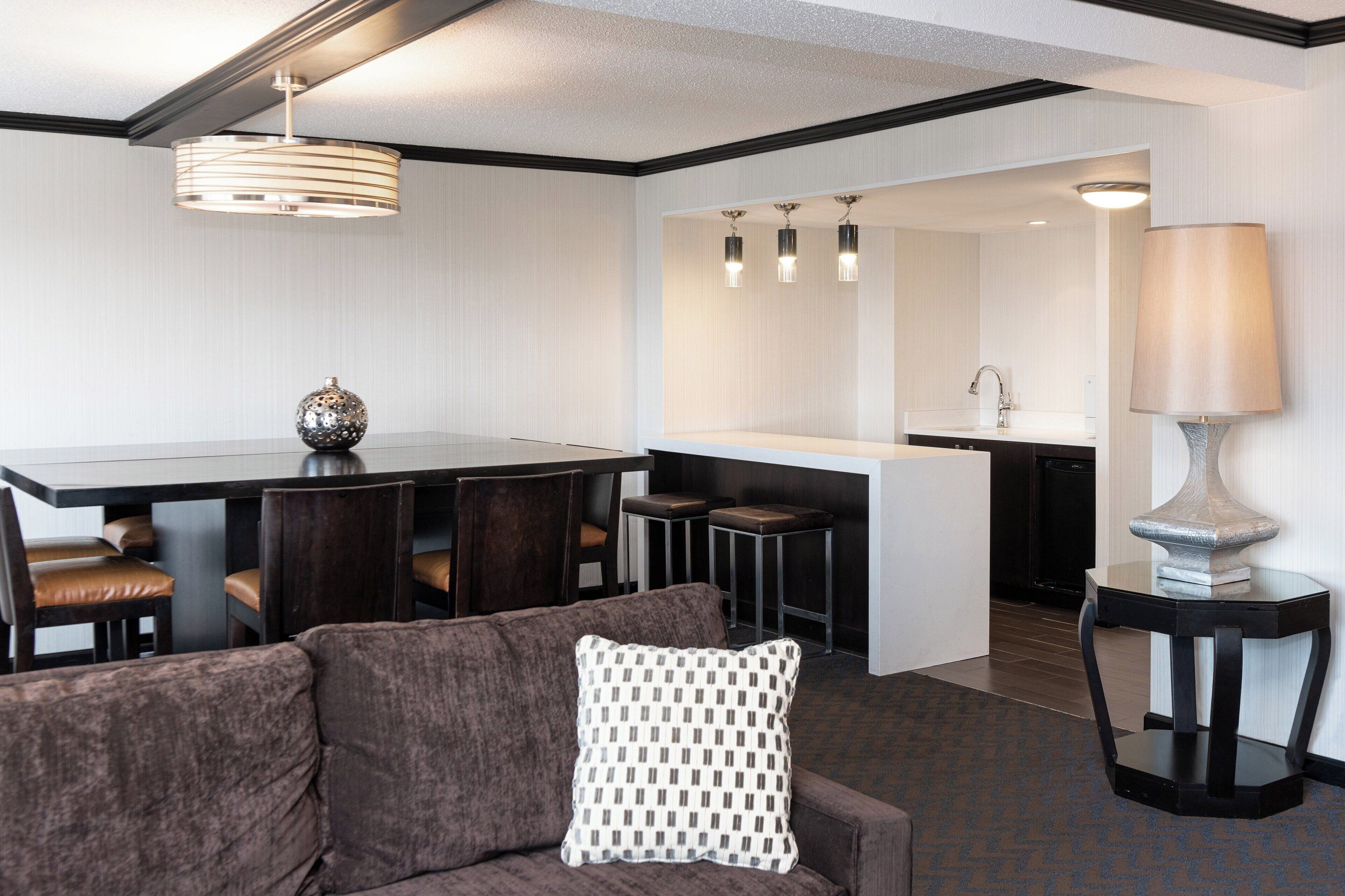 A modern hotel room with a dark brown sofa, a patterned throw pillow, a dining area with a table and chairs, a kitchen with a sink, and a lamp on a side table.