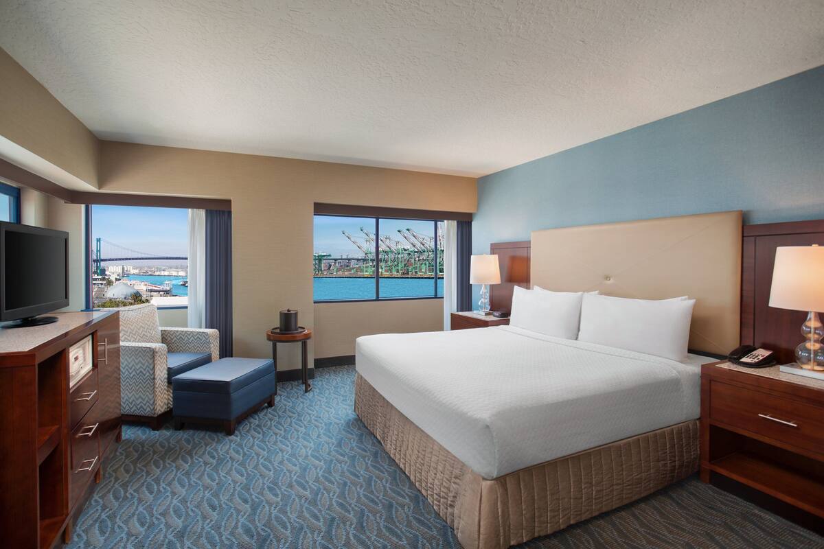 A hotel room with a large bed, a television, a desk with a chair, and a view of a bridge and water.