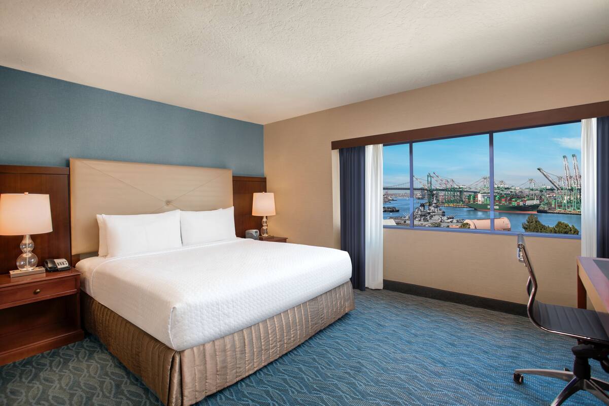 A hotel room with a large bed, two bedside tables with lamps, a desk with a chair, and a large window with a view of a cityscape and water.