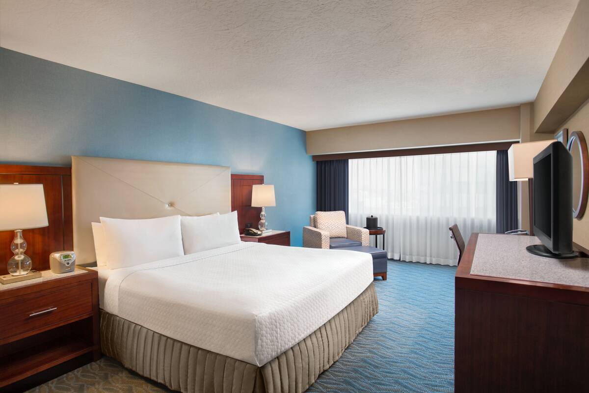 A hotel room with a large bed, a desk with a television, a chair, and a desk lamp.