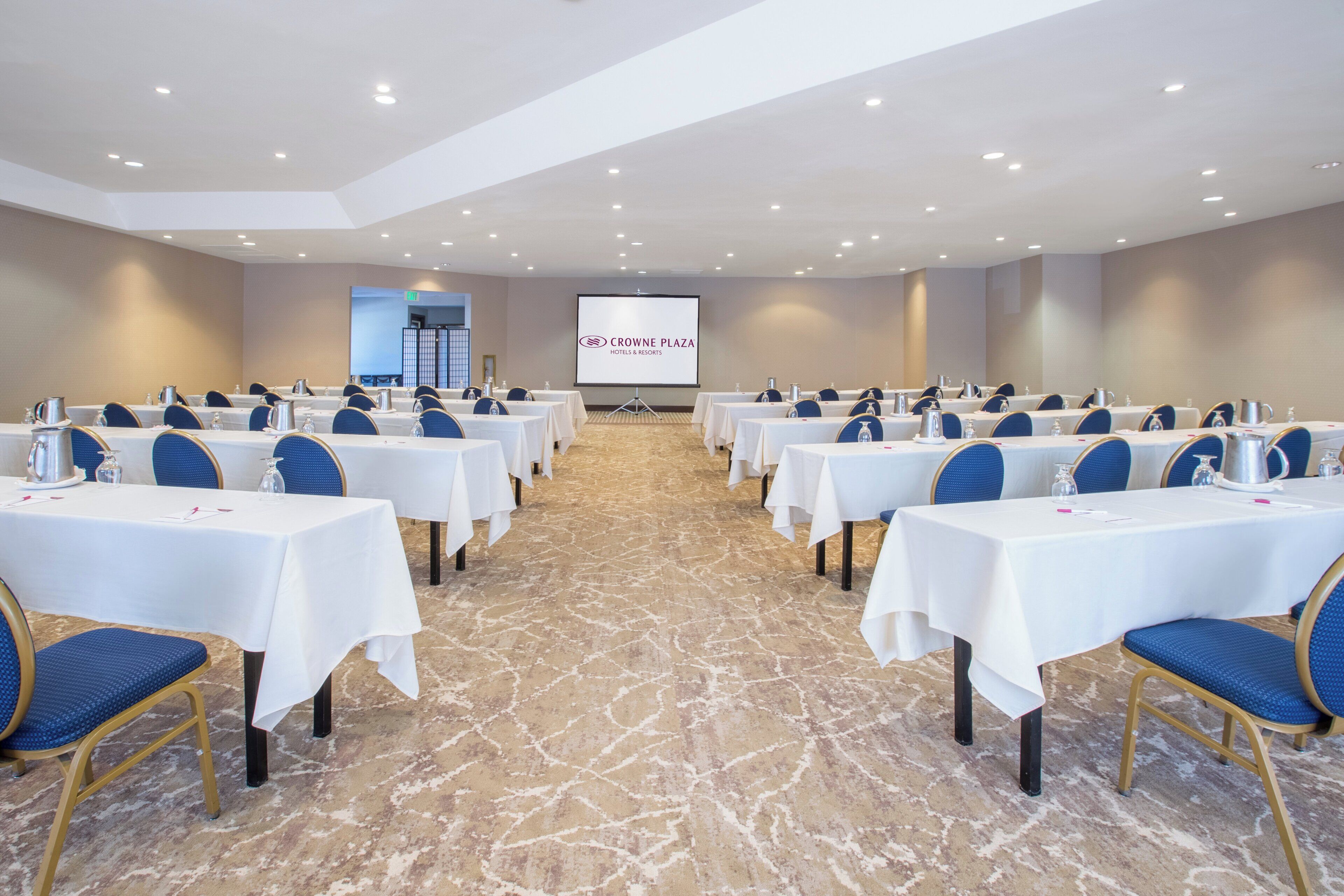 A conference room with rows of tables, chairs, and a projection screen in the background.
