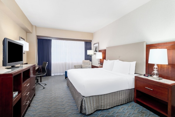 A hotel room with a large bed, a desk with a television, a chair, a nightstand with a lamp, and a window with curtains.