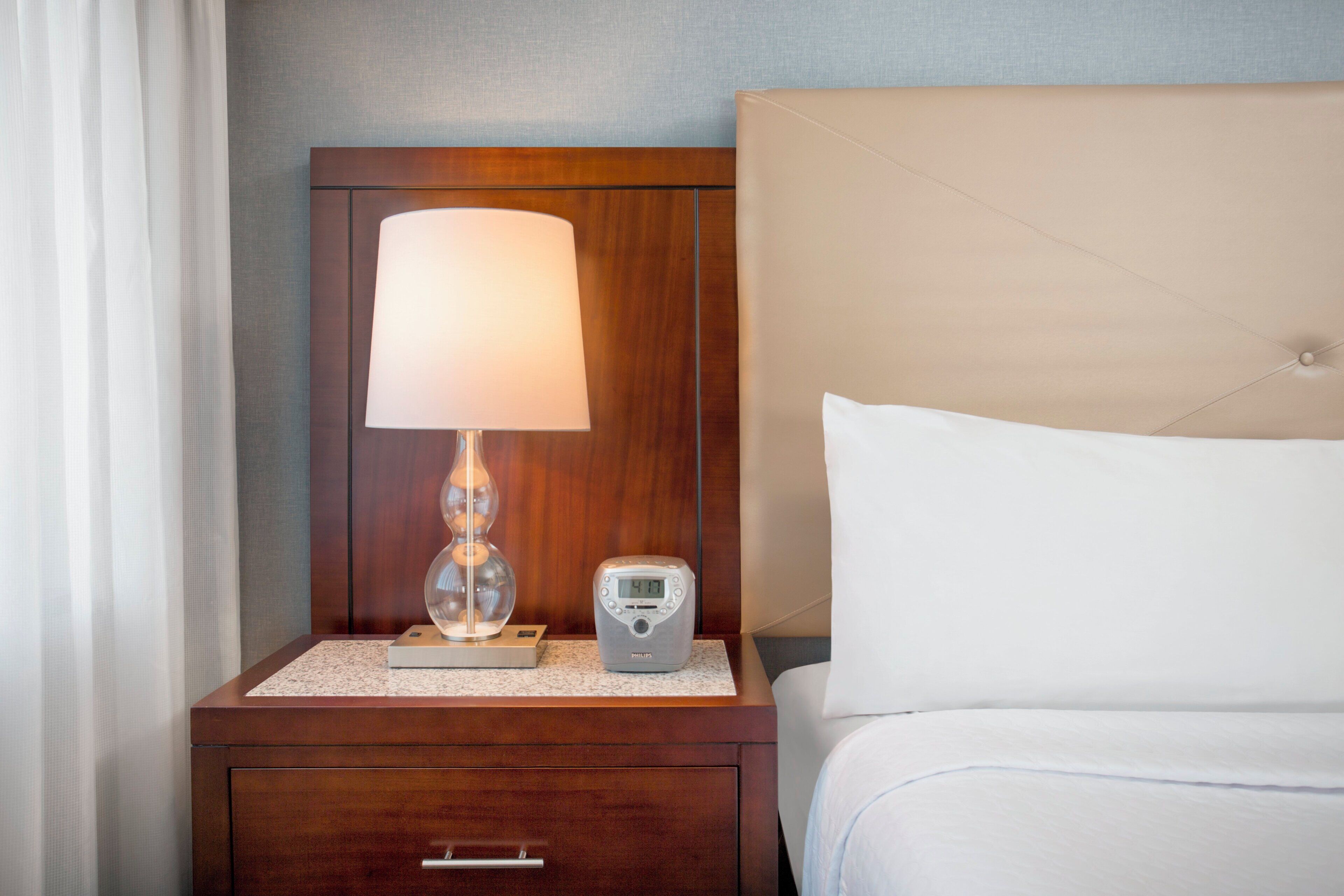 Une chambre d’hôtel avec une table de chevet en bois, une lampe, une horloge numérique et un lit avec une literie blanche.