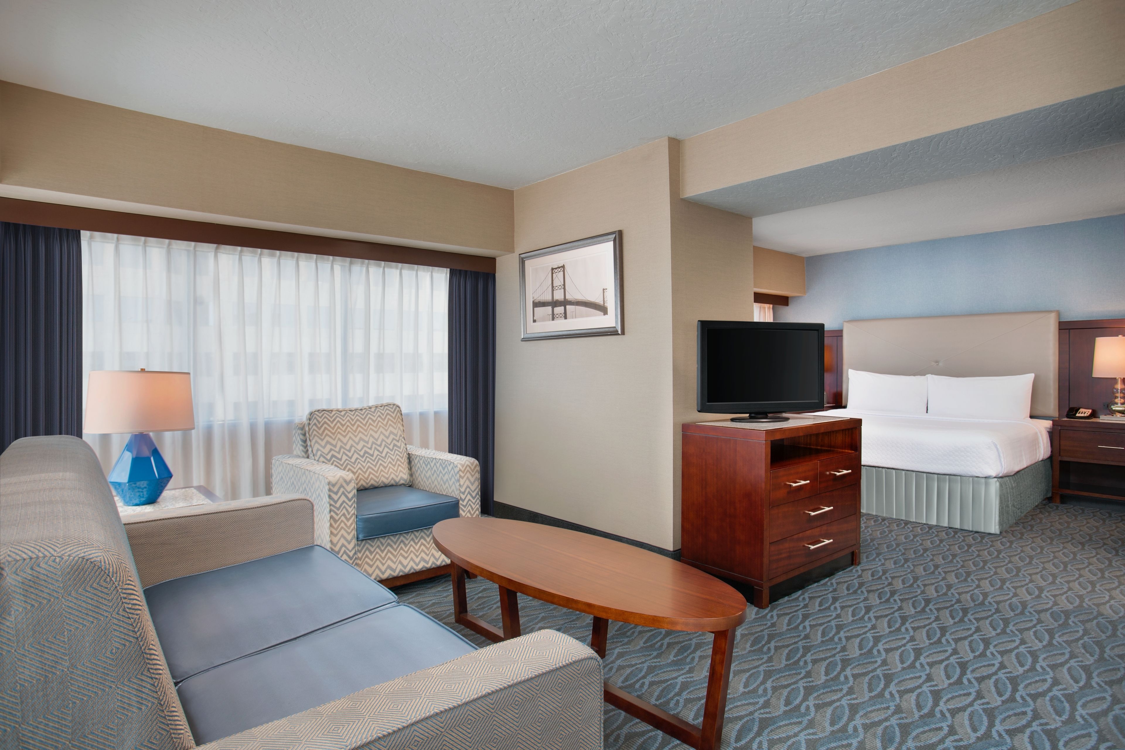 A hotel room with a sofa, armchair, TV, bed, and a desk with drawers.