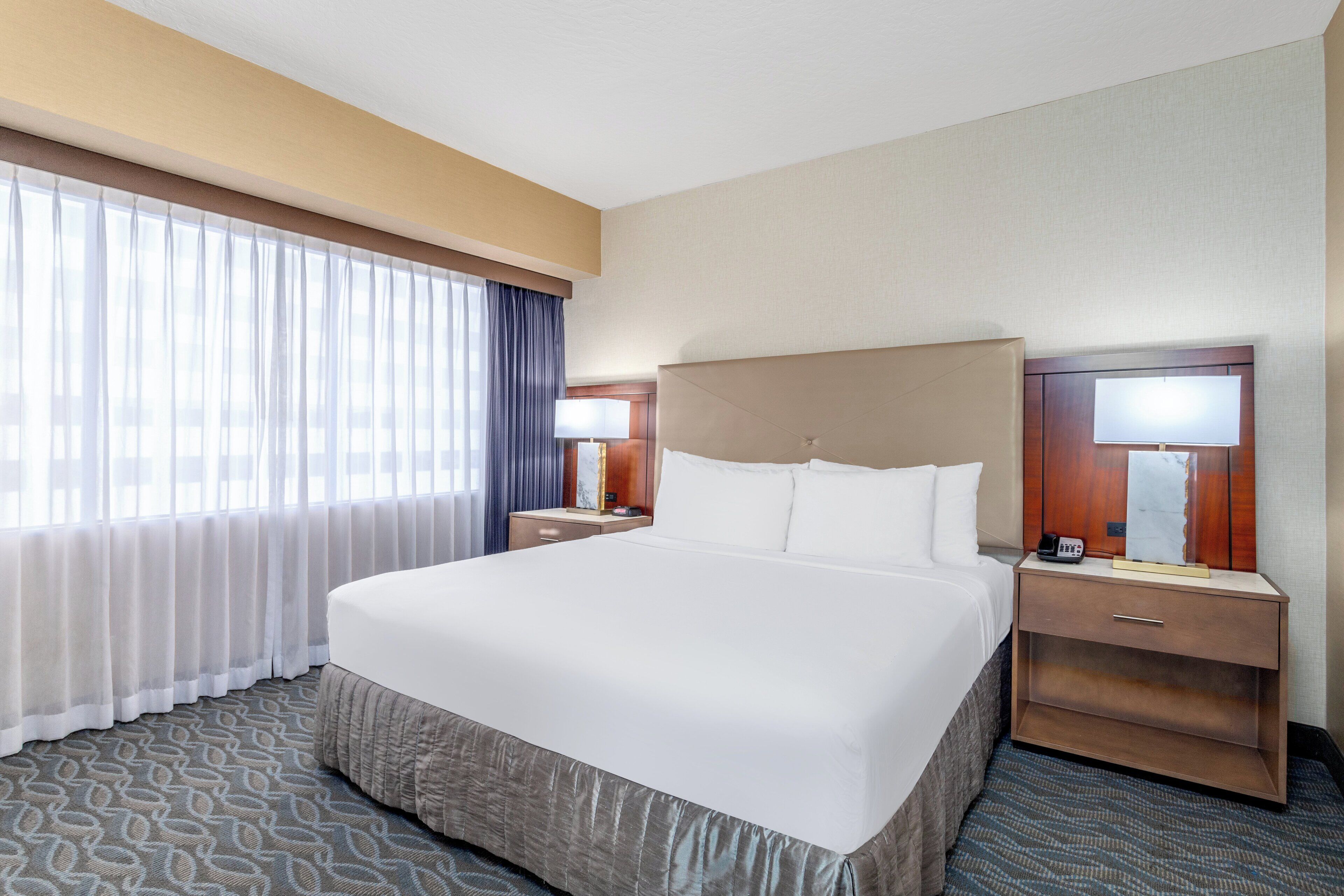 A hotel room with a large bed, a nightstand with a lamp, and a window with curtains.