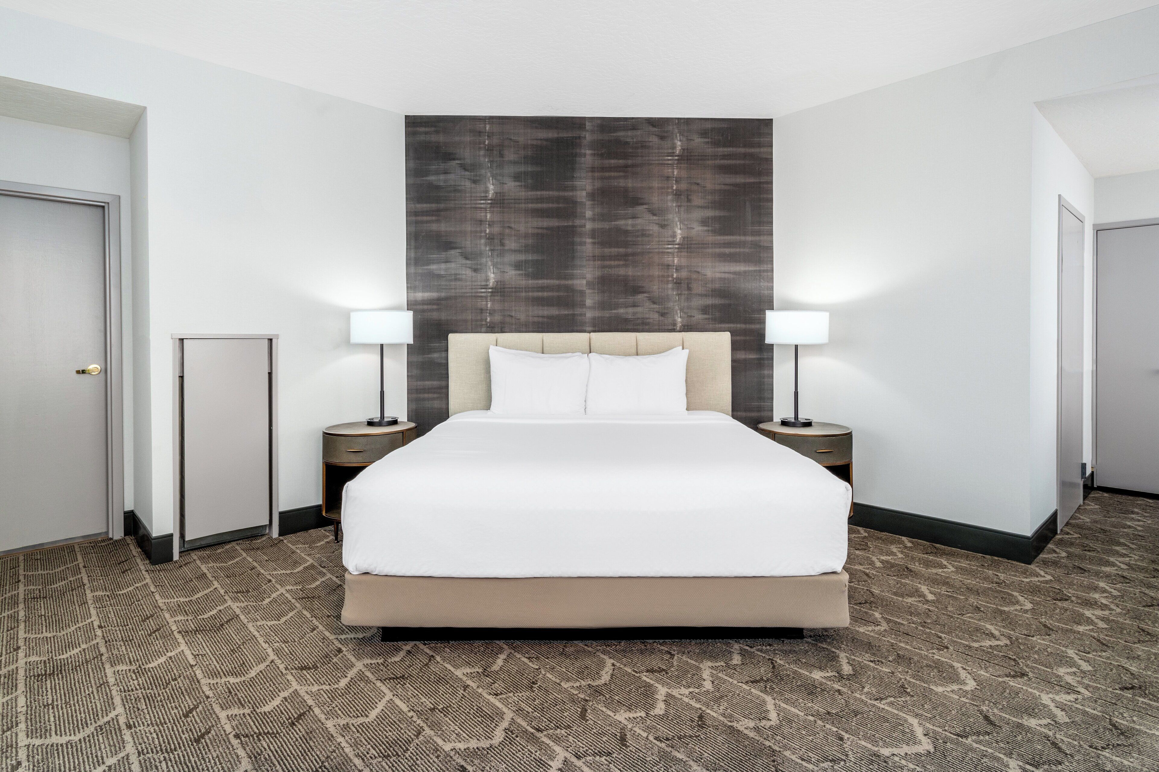 Habitación de hotel con una cama grande, dos mesitas de noche con lámparas, cabecero y suelo alfombrado.