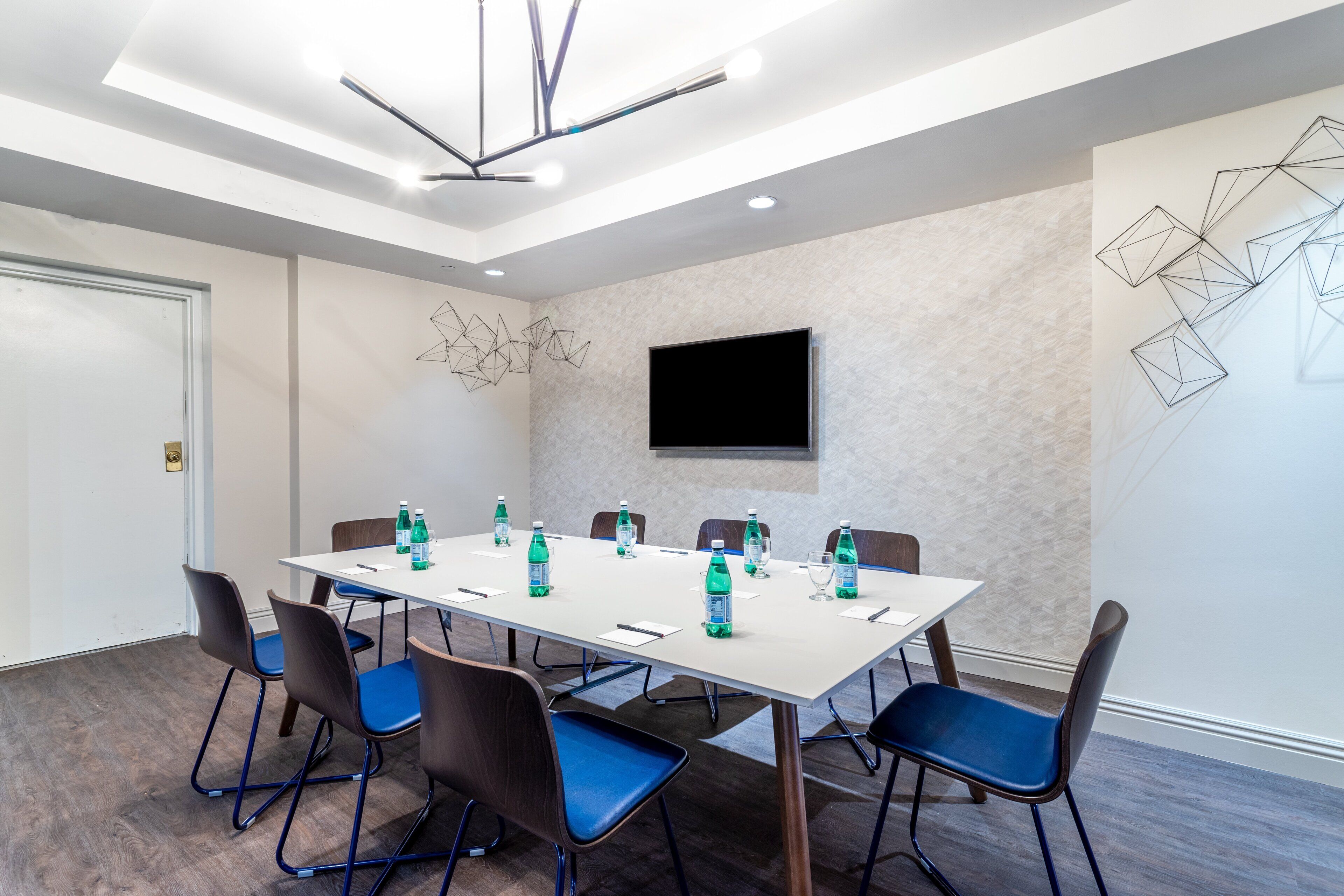 A conference room with a long table, blue chairs, water bottles, and a flat-screen TV on the wall.
