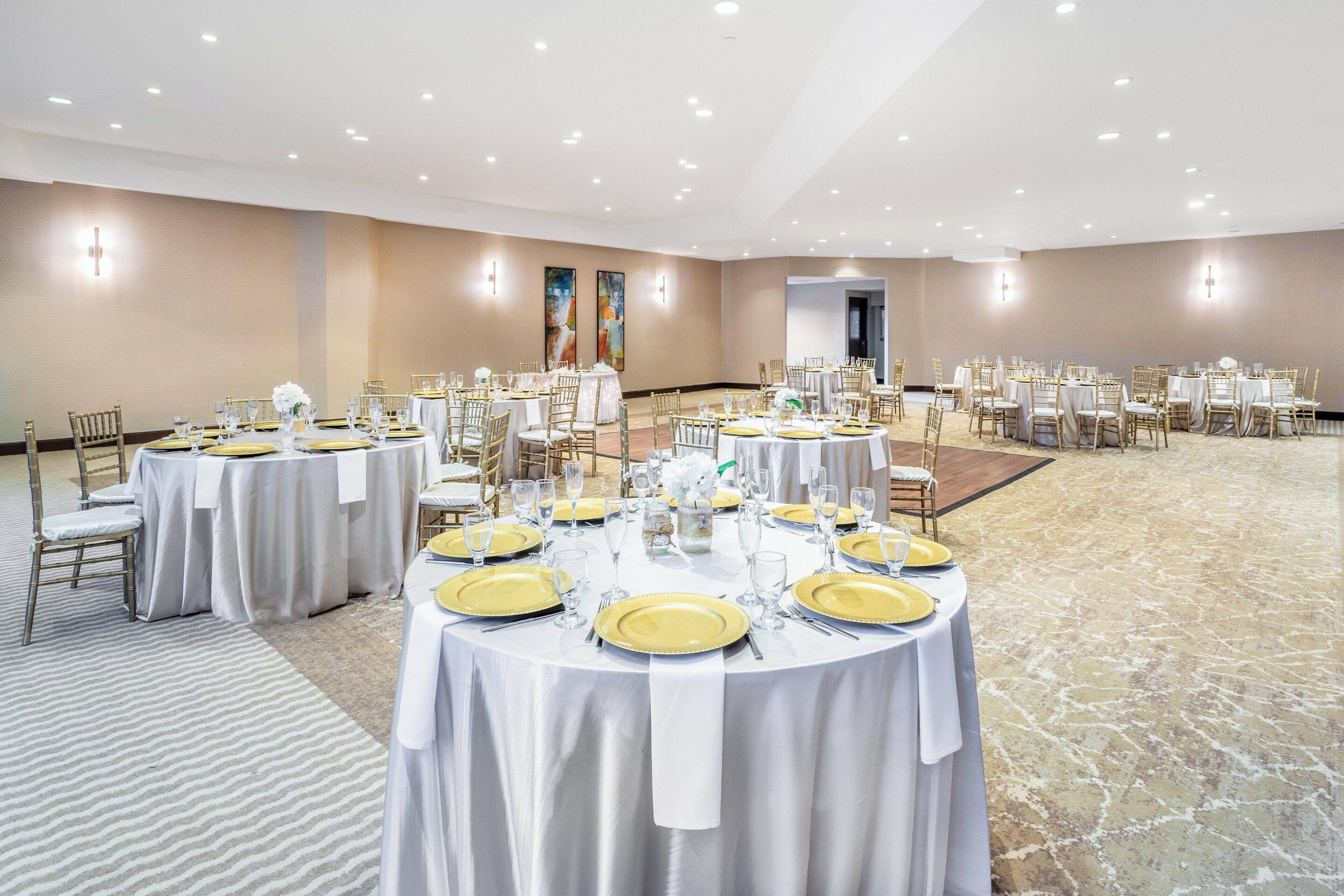 A large conference room with multiple round tables set for dining, featuring white tablecloths and gold accents.