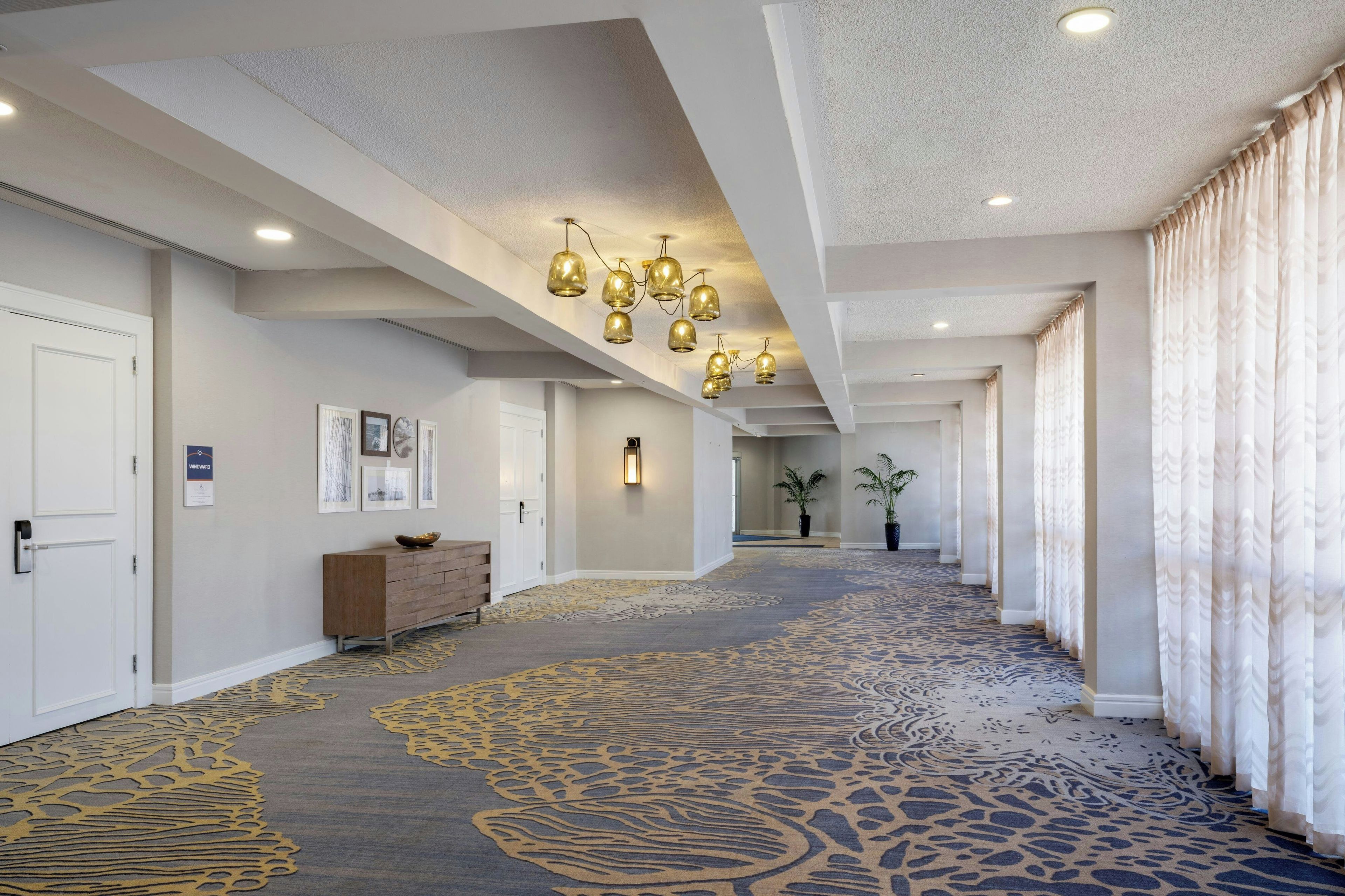 A hotel corridor with a patterned carpet, a wooden bench, a wall-mounted light fixture, and a door on the left.