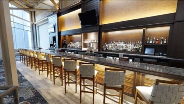A bar area with a long counter, multiple bar stools, and shelves with bottles. - Southbank Hotel by Marriott Jacksonville Riverwalk (Jacksonville)