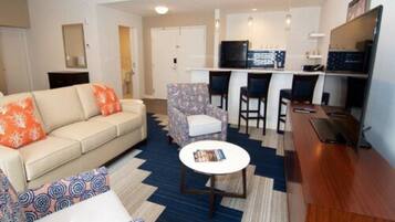 A modern living room with a sofa, patterned armchair, and a small coffee table. There is a kitchen area with a counter and bar stools.