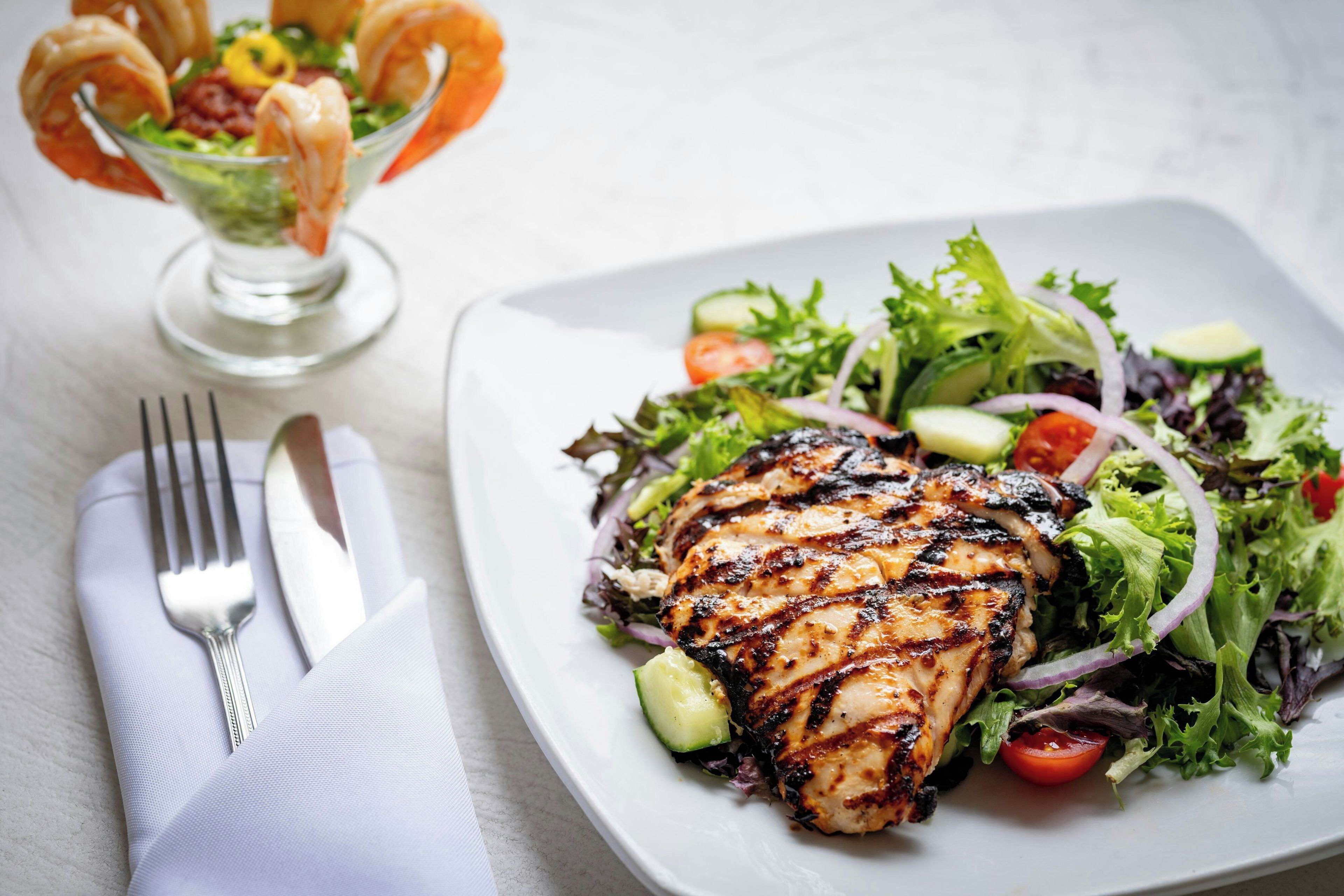 A plate of grilled chicken with a side salad and a glass of a seafood cocktail.