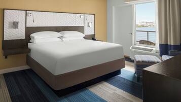 A hotel room with a large bed, a desk, a chair, a window with a view of the water, and a door.