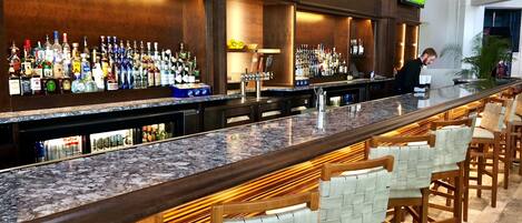 A bar area with a long counter, multiple seating options, and a television screen displaying a sports event.