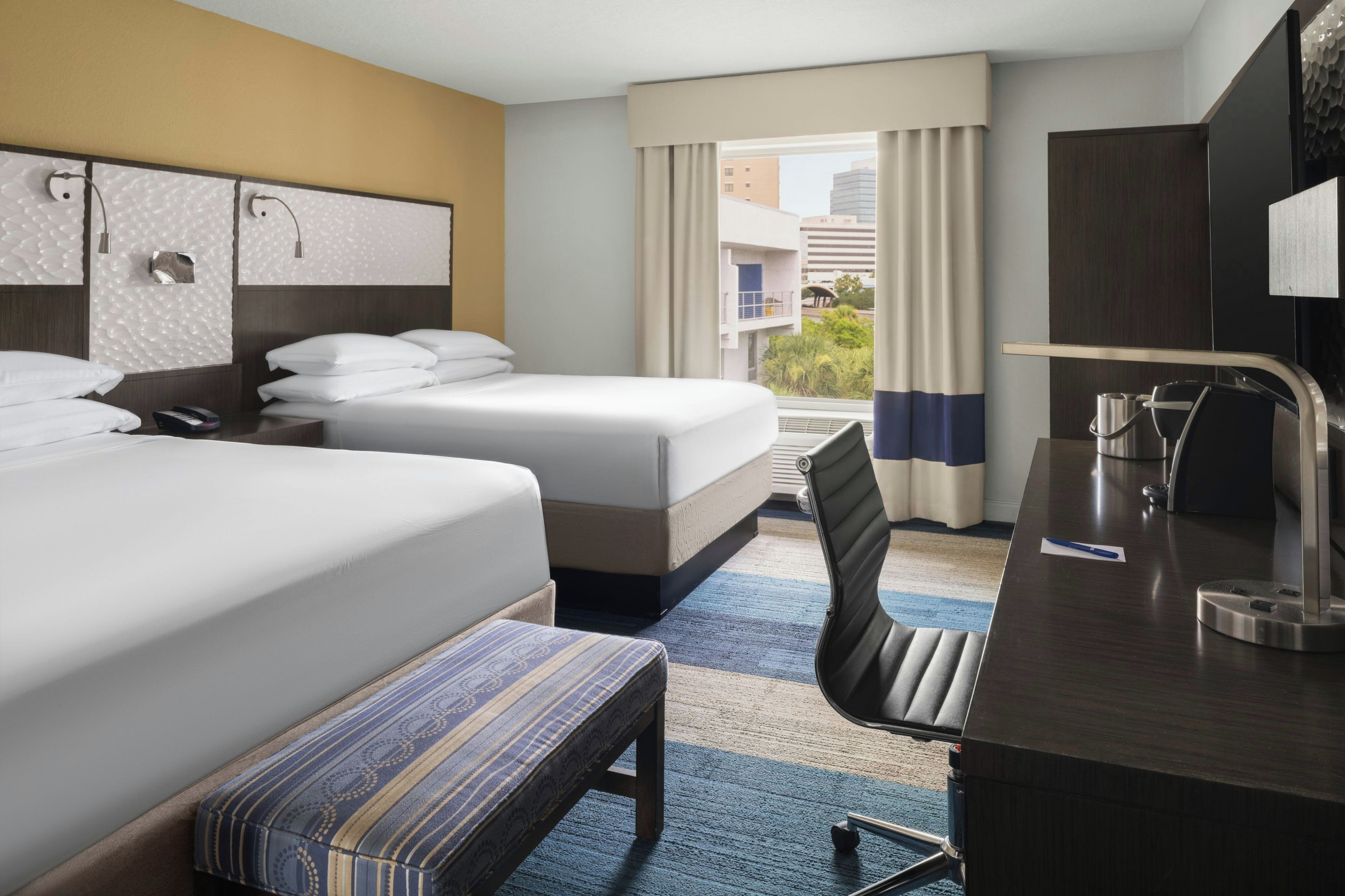 A hotel room with two beds, a desk, a chair, and a view of the city.