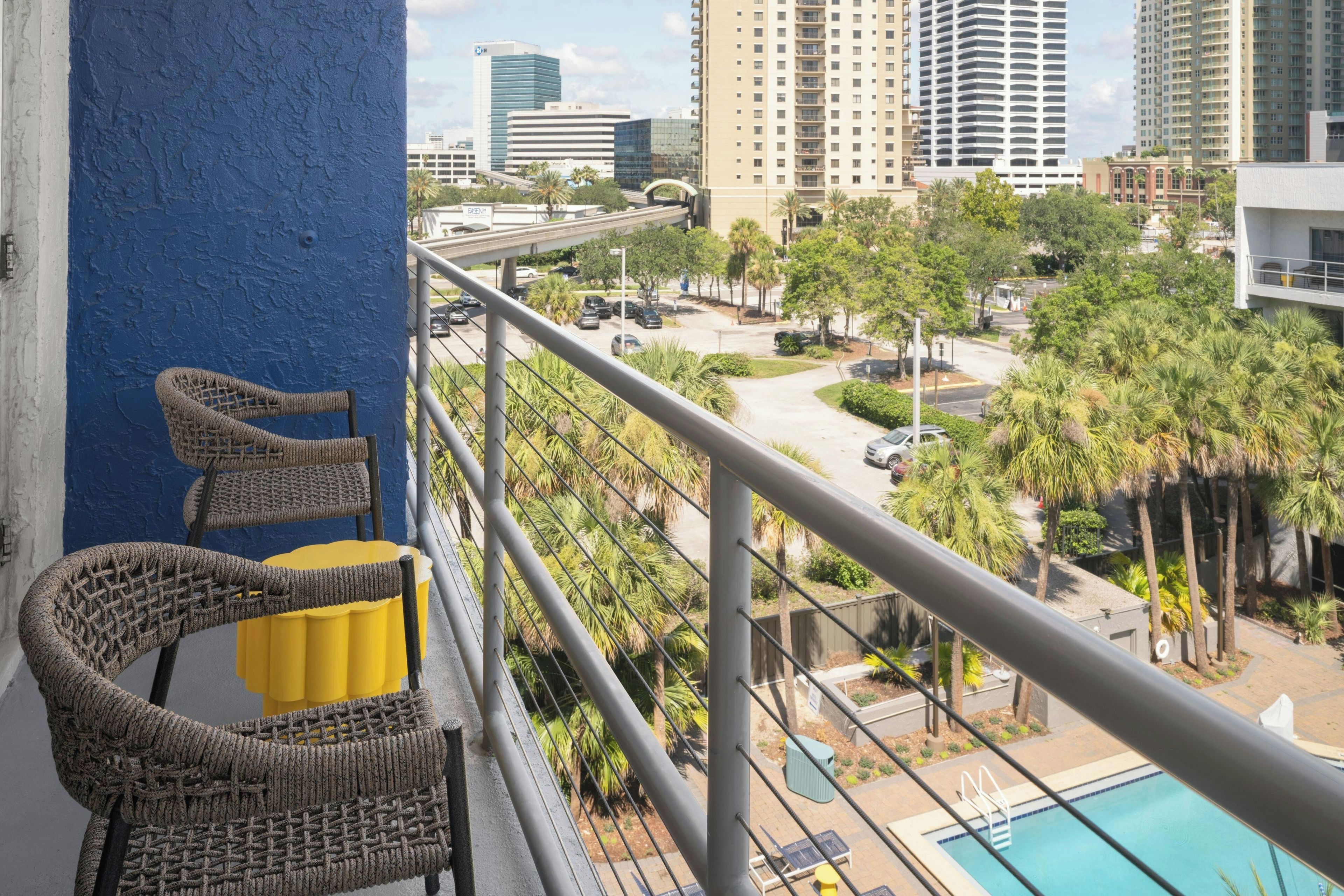 A balcony with wicker chairs and a yellow cushion, overlooking a swimming pool and a parking area with palm trees.