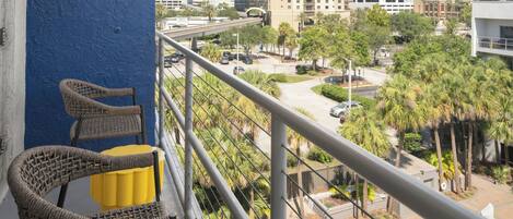A balcony with wicker chairs and a yellow cushion, overlooking a swimming pool and a parking area with palm trees.