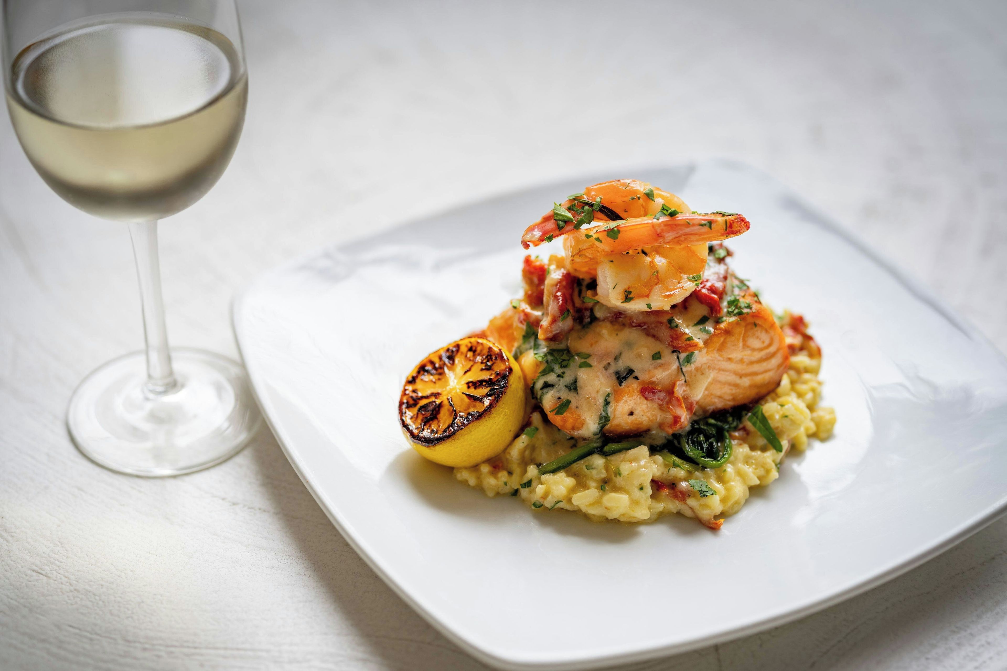A square plate with a seafood dish, a glass of white wine, and a slice of grilled lemon on a white table.