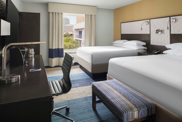 A hotel room with a desk, two beds, a chair, and a view of buildings.