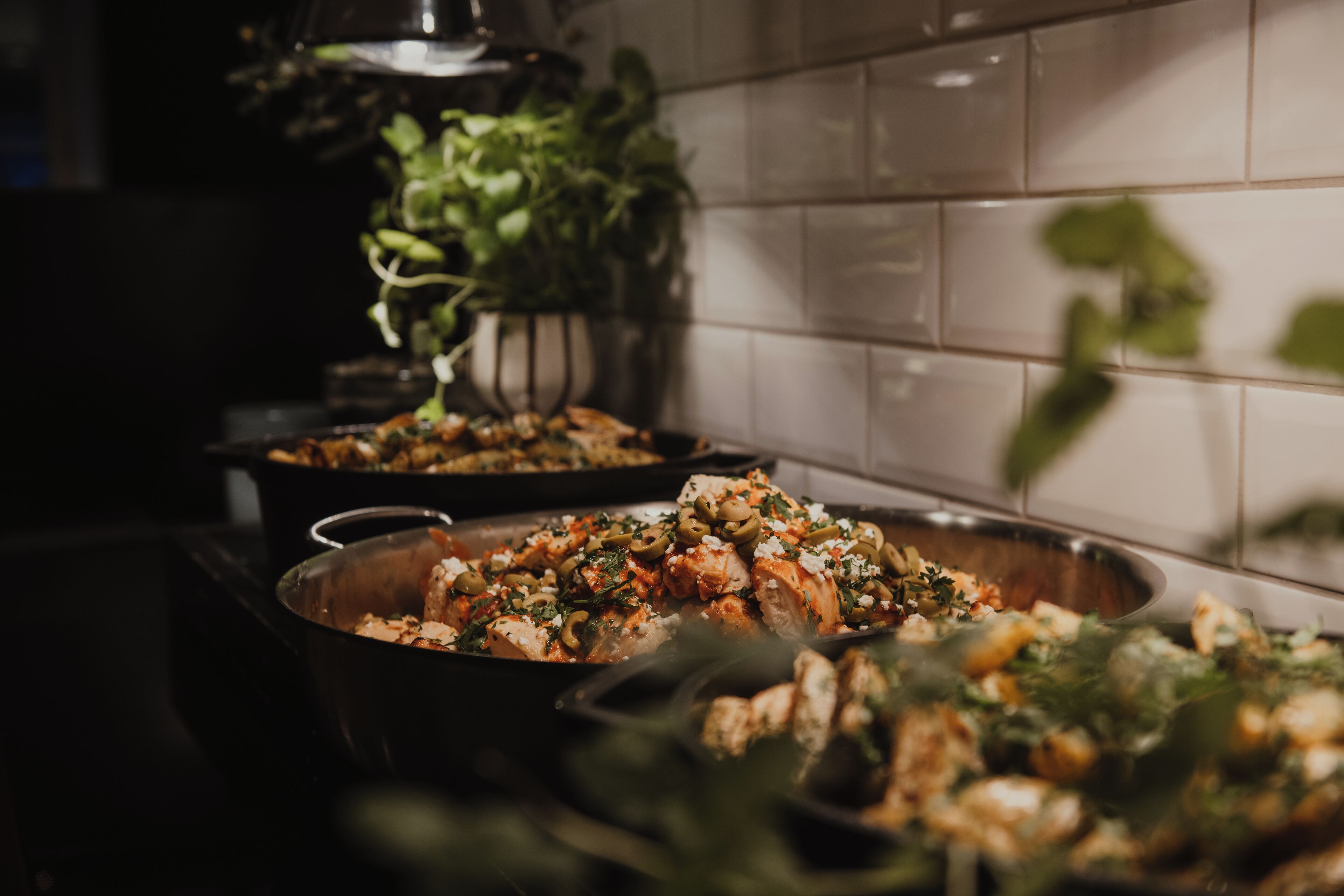 A kitchen with two large pans of food, a plant, and a tiled backsplash.