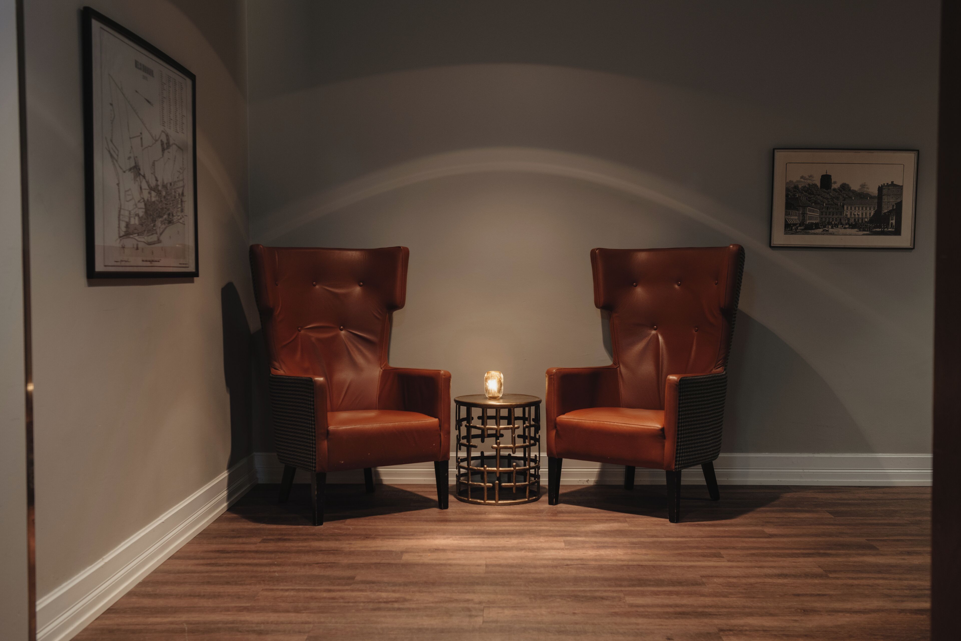 Two leather armchairs facing each other with a small round table between them, a lit candle on the table, and framed artwork on the walls.