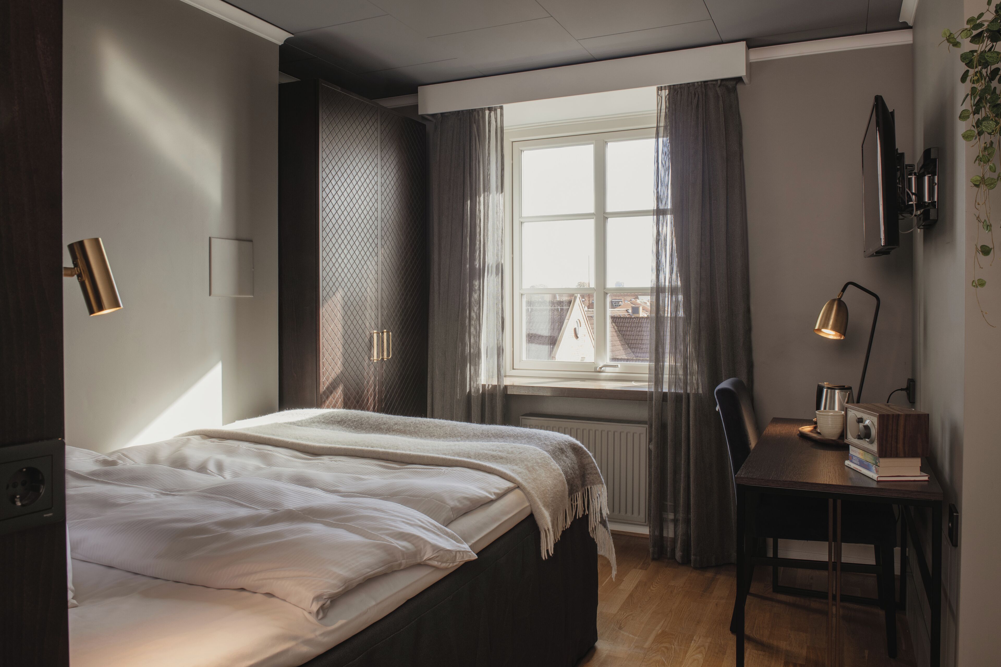 Un dormitorio con cama, escritorio, silla, ventana con cortinas y lámpara de pared.