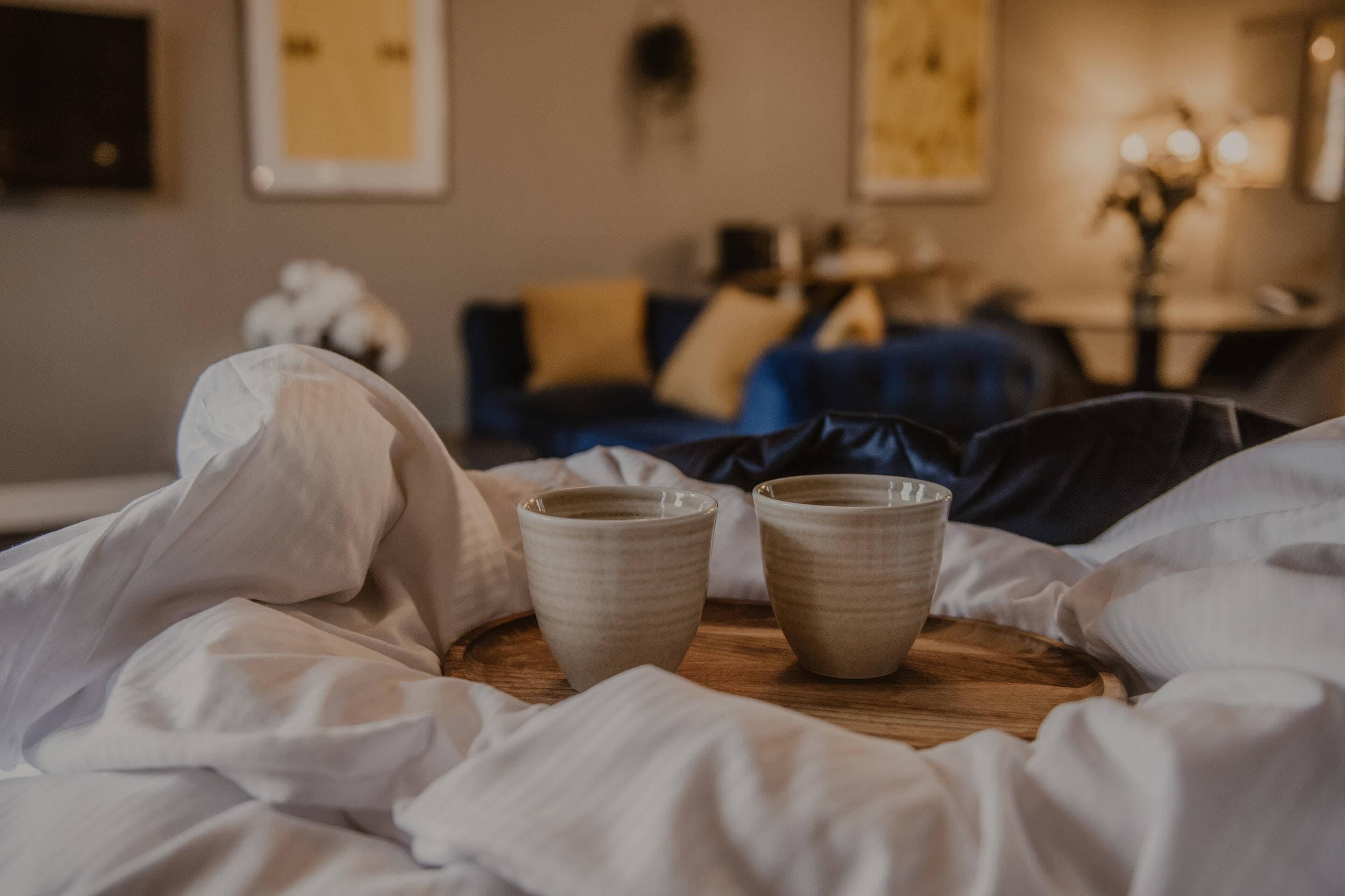 A bed with white bedding and two ceramic cups on a wooden tray.