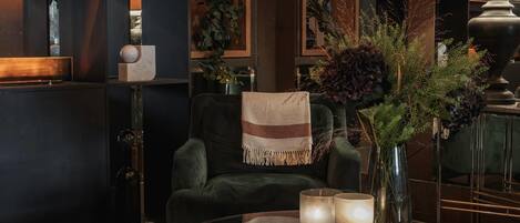A cozy lounge area with a dark green armchair, a glass coffee table with candles, and a vase with dried plants.