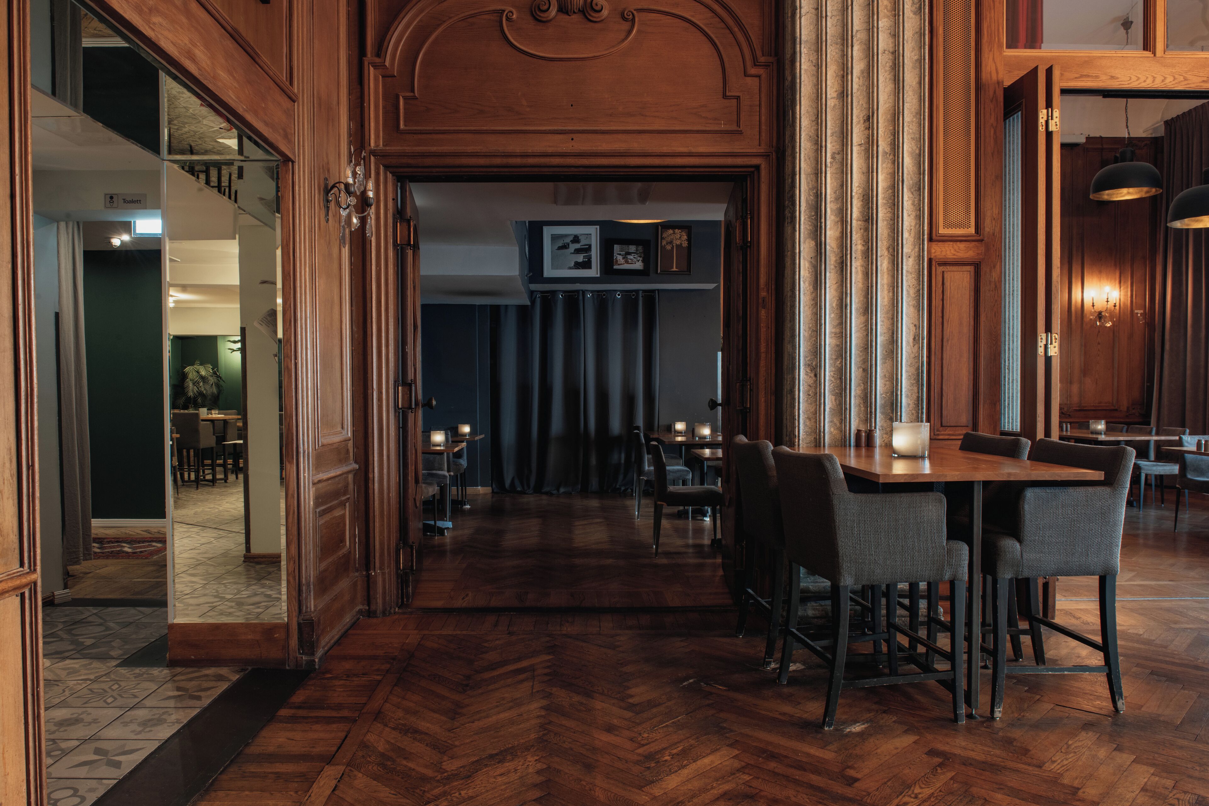 A wooden interior with a herringbone floor, a bar area with bar stools, and a dining area with tables and chairs.