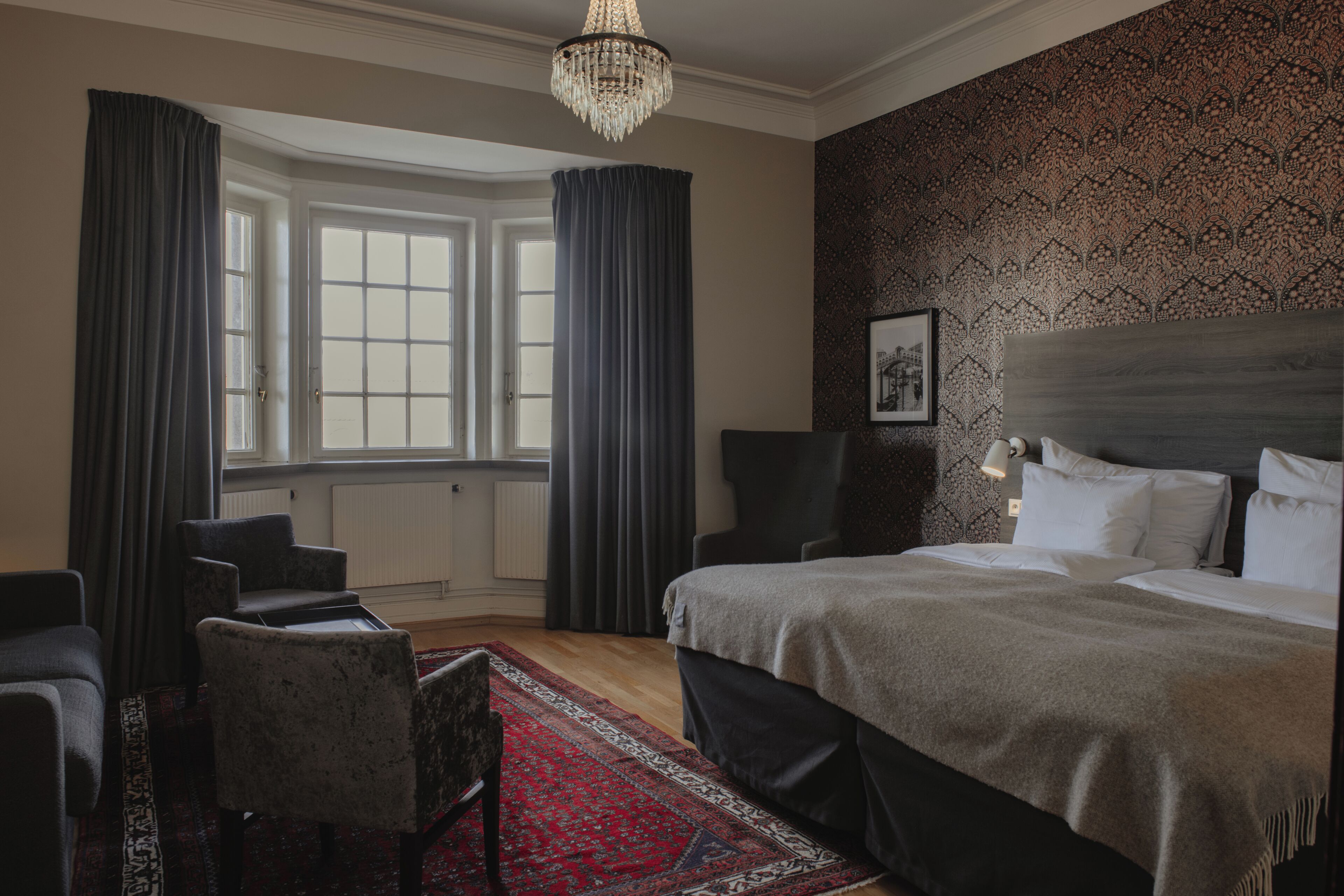Ett hotellrum med en stor säng, två fåtöljer, en röd, mönstrad matta, en kristallkrona och ett fönster med gardiner.