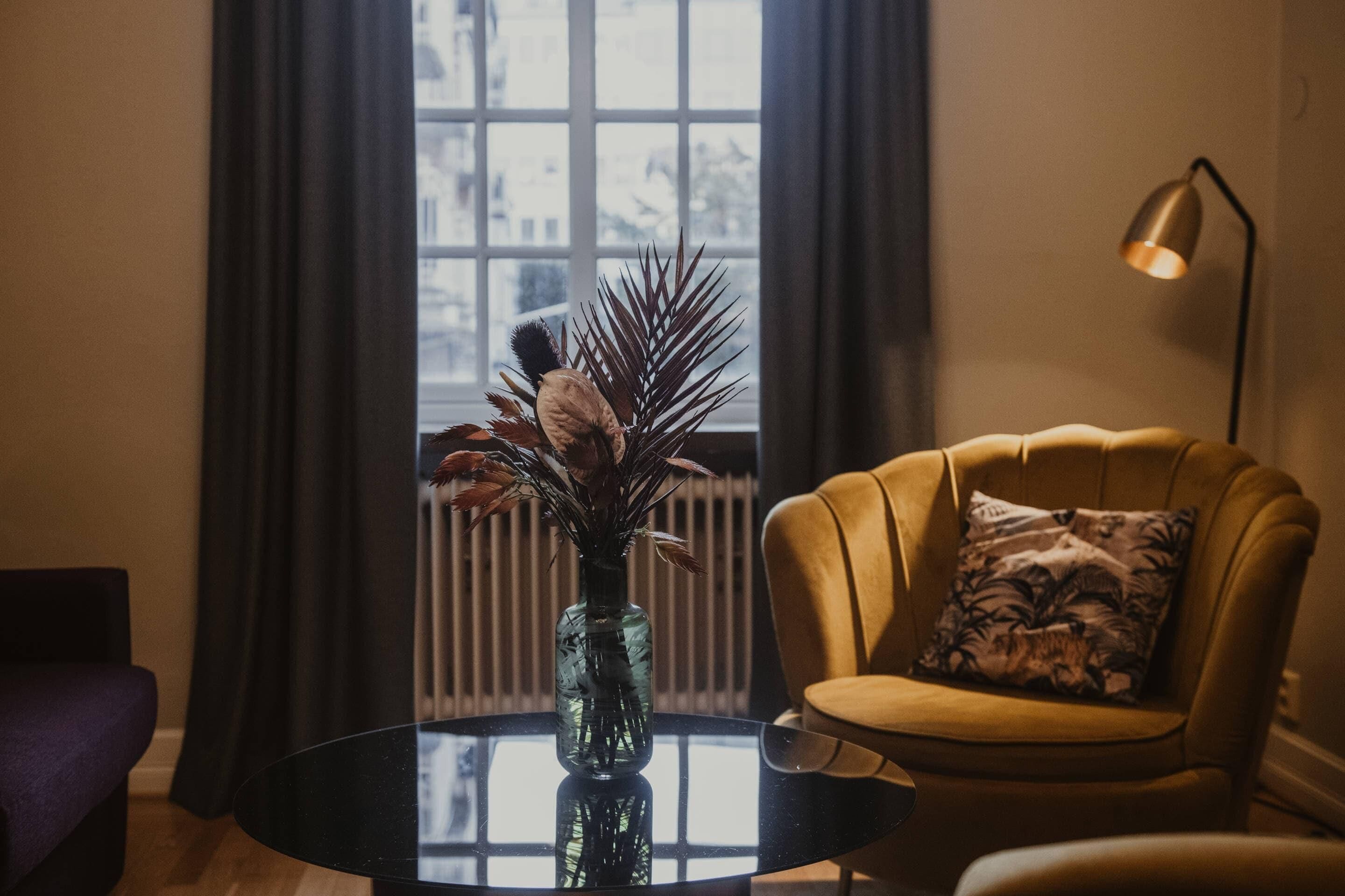 Una habitación con una mesa redonda de vidrio, un sillón amarillo con un cojín estampado y un jarrón con plantas secas.
