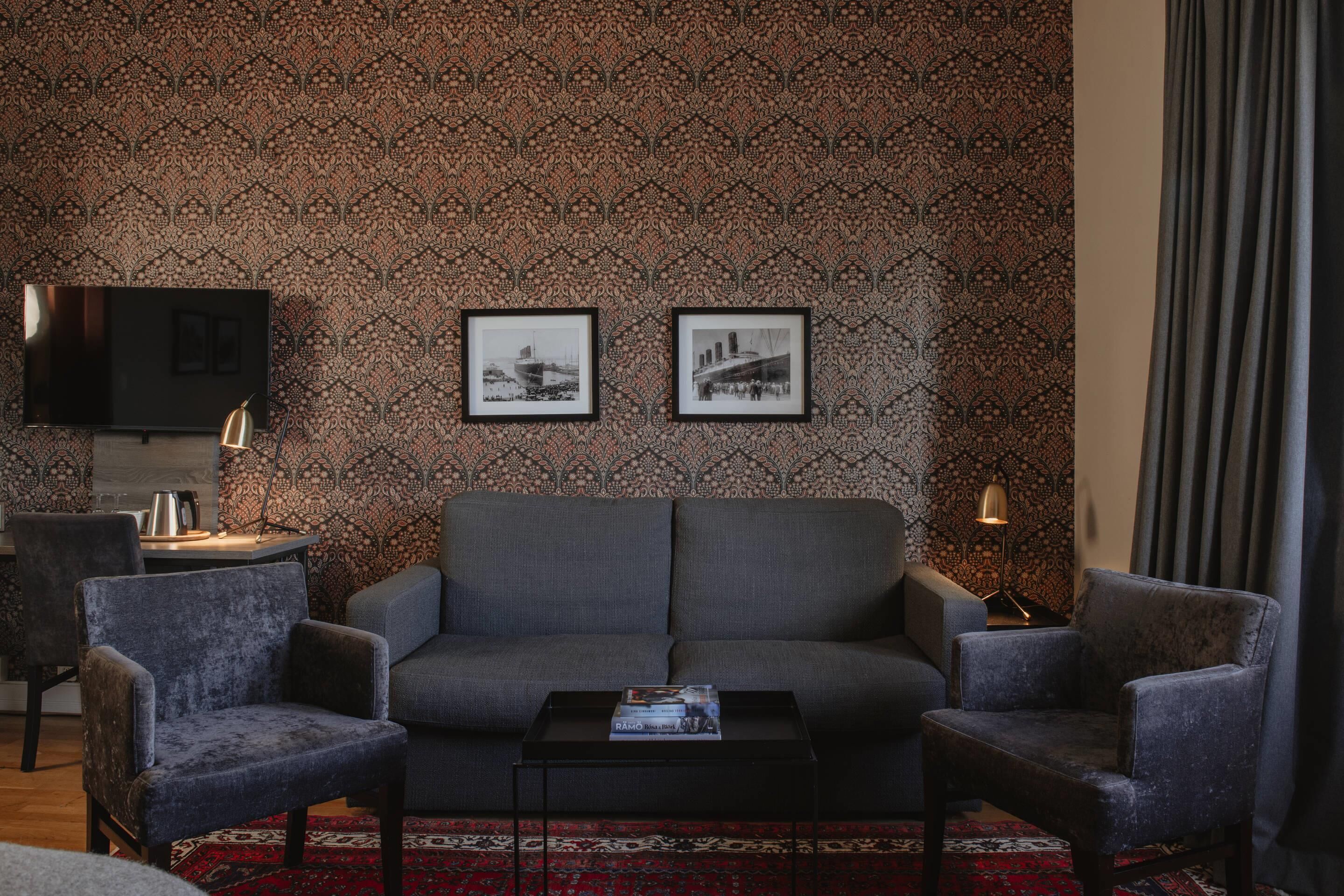 A living room with a patterned wallpaper, a grey sofa, two armchairs, a coffee table, and a flat-screen TV mounted on the wall.