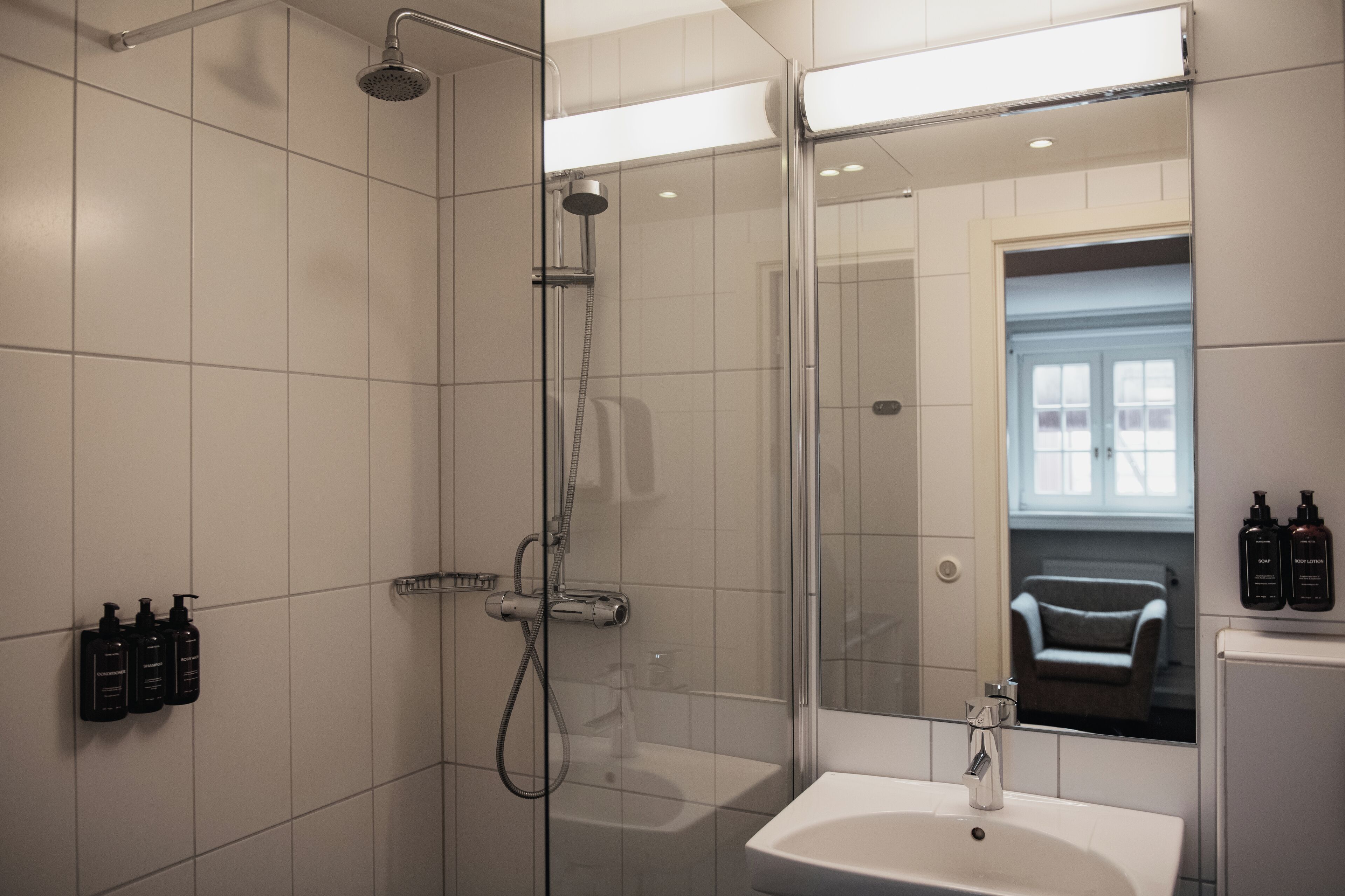A modern bathroom with a glass-enclosed shower, a white sink, and a mirror reflecting a chair and window.
