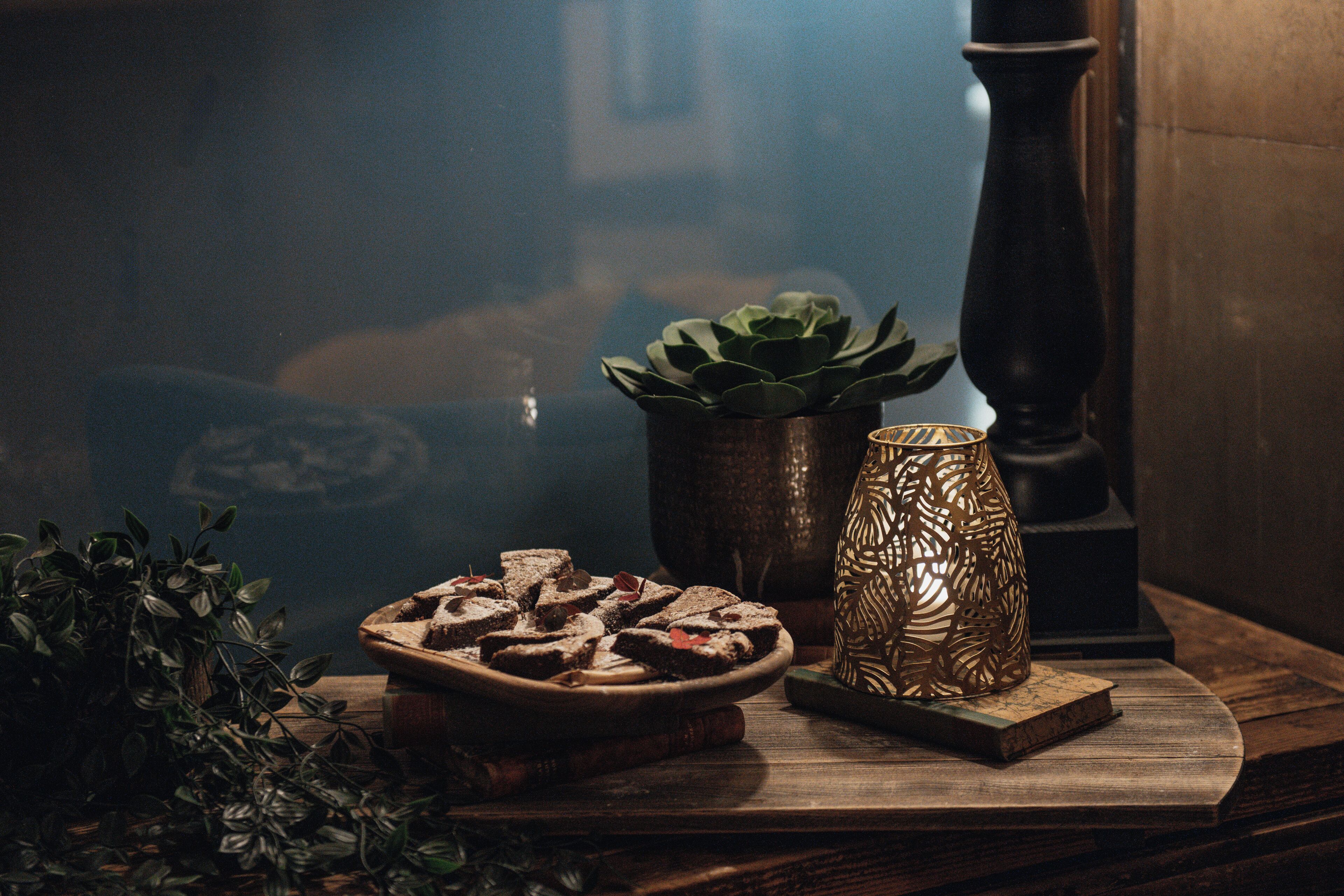Una mesa de madera con un plato de brownies, una suculenta en maceta y un candelabro decorativo.