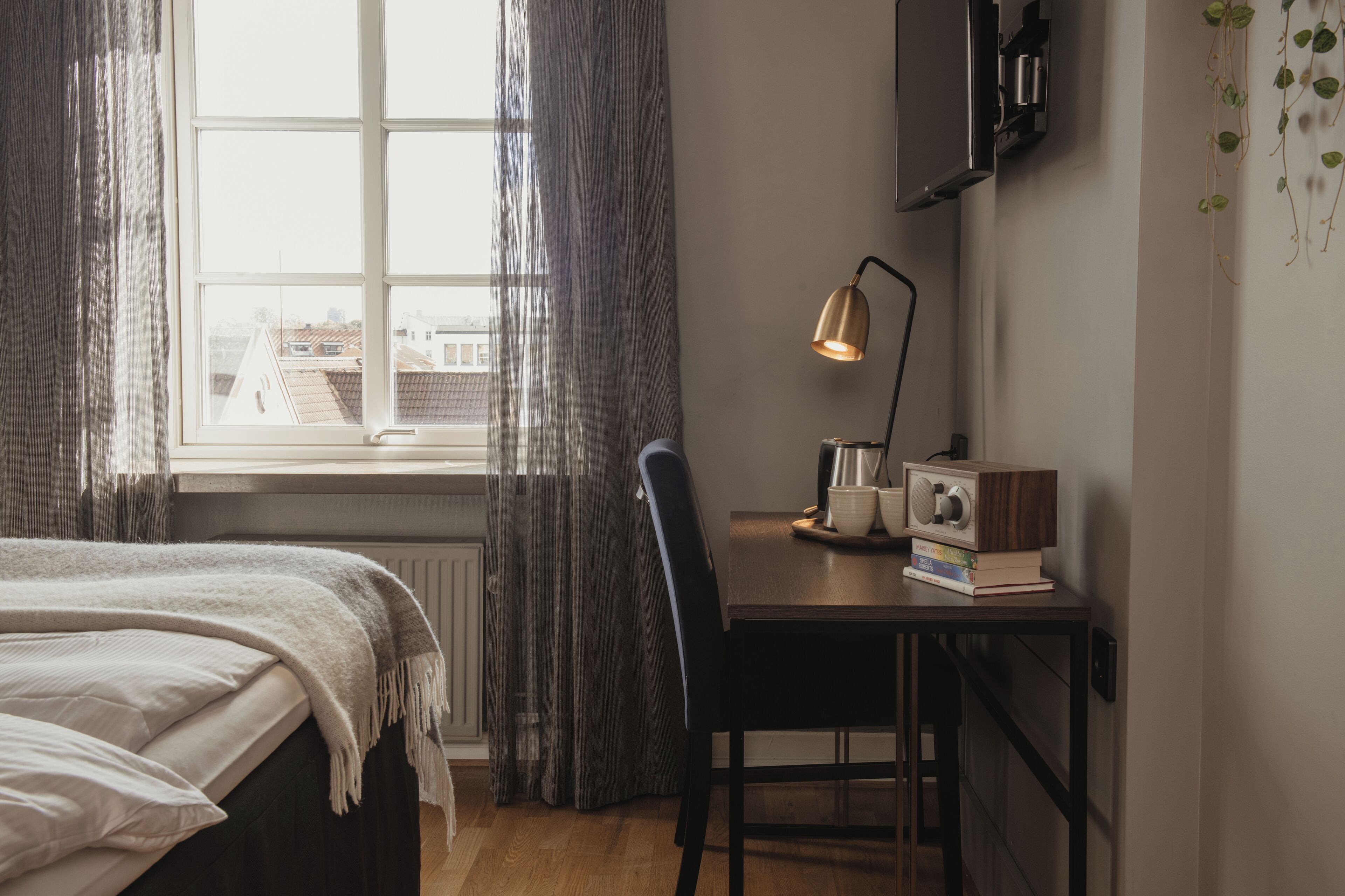 Un dormitorio con cama, escritorio con lámpara, radio y vista a edificios a través de una ventana.