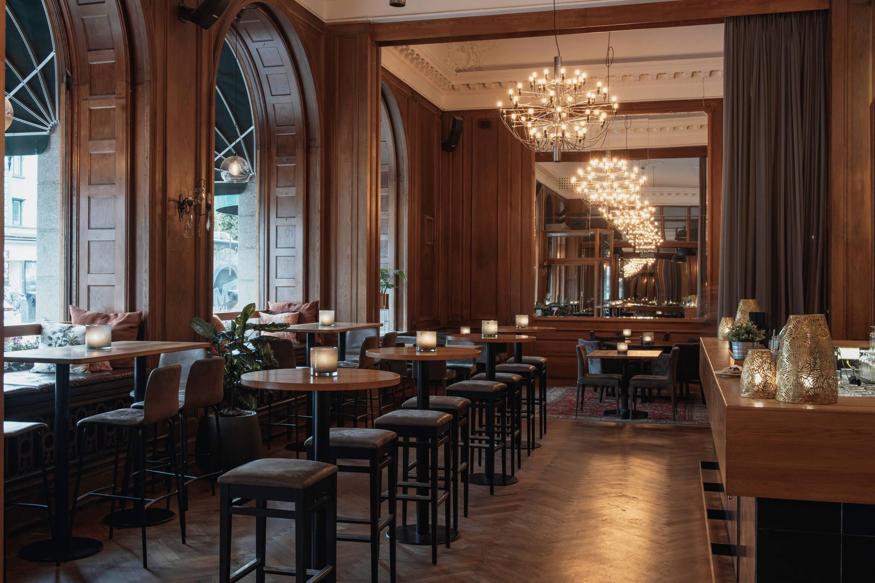 Restaurante com painéis de madeira, amplas janelas em arco e um candelabro. Mesas dispostas com velas e banquetas.