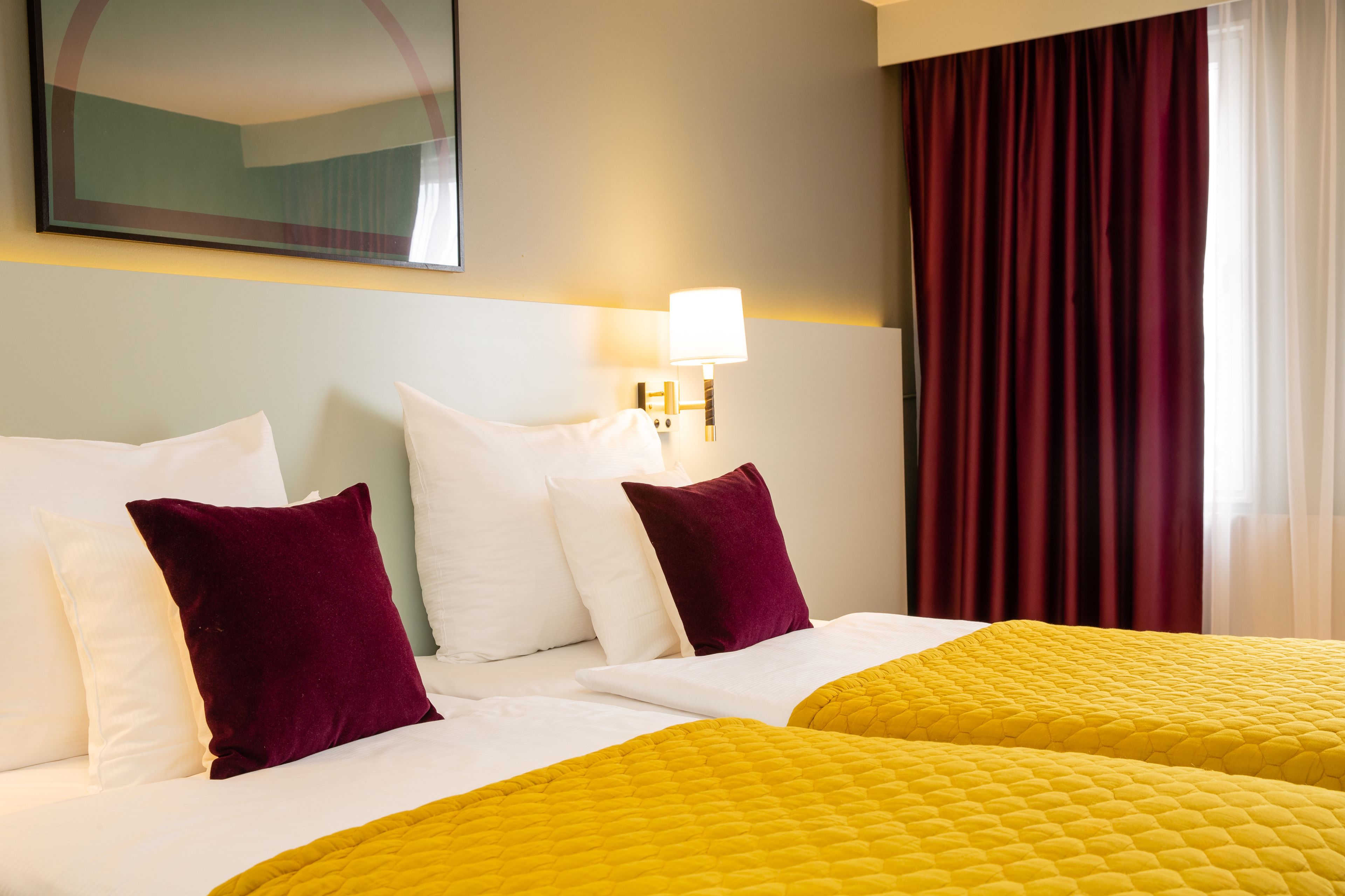 Habitación de hotel con dos camas, ropa de cama blanca y amarilla, una cortina roja y una lámpara fijada a la pared.