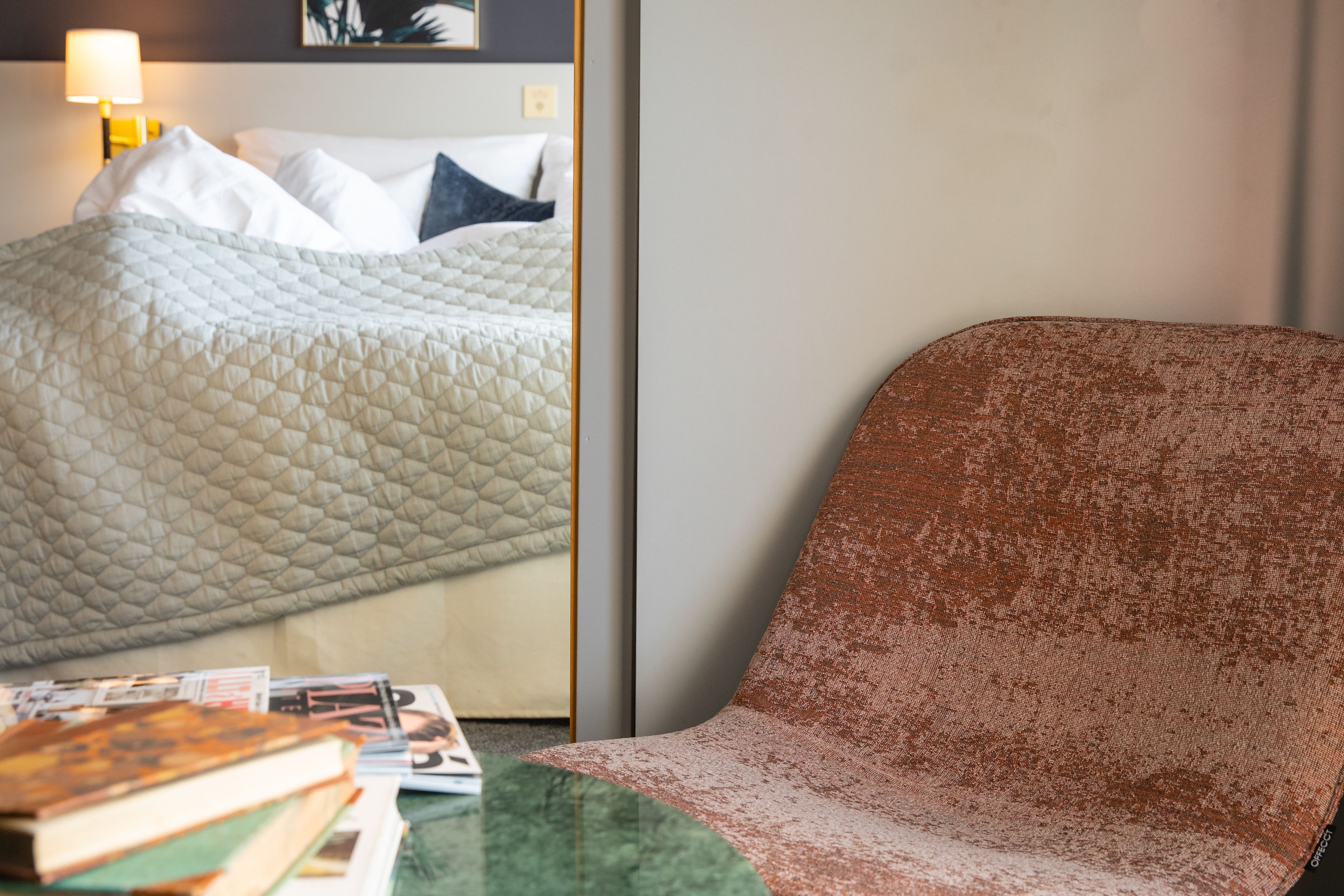 A hotel room with a bed, pillows, a green table with books, and a brown armchair.