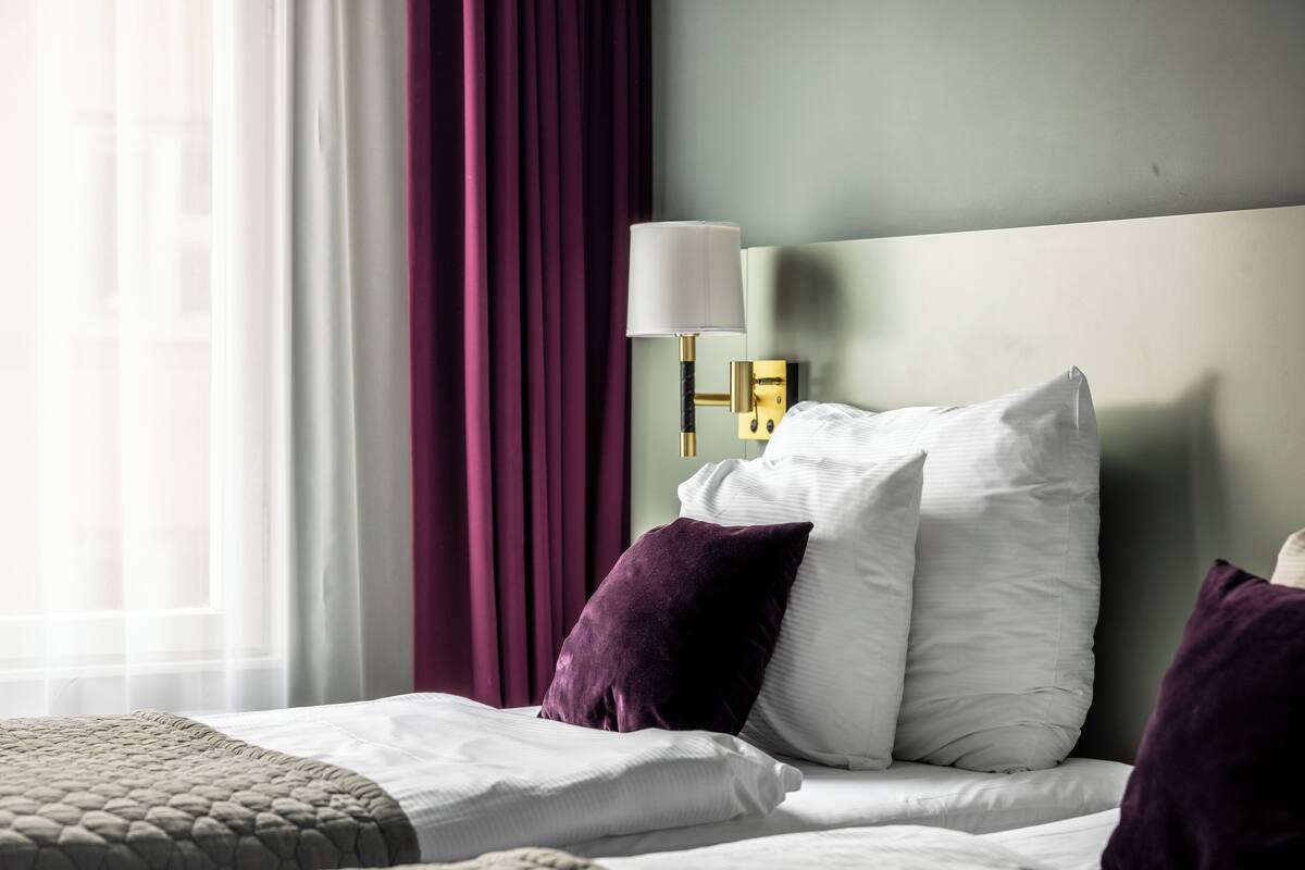 Un lit bien fait, avec des oreillers blancs et violets, une lampe de chevet dorée et une vue sur une fenêtre avec des rideaux.