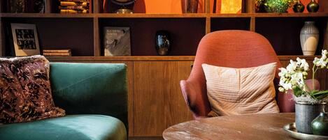 A cozy living room with a green sofa, a wooden coffee table, and a red armchair. Shelves above contain books, a globe, and decorative items.