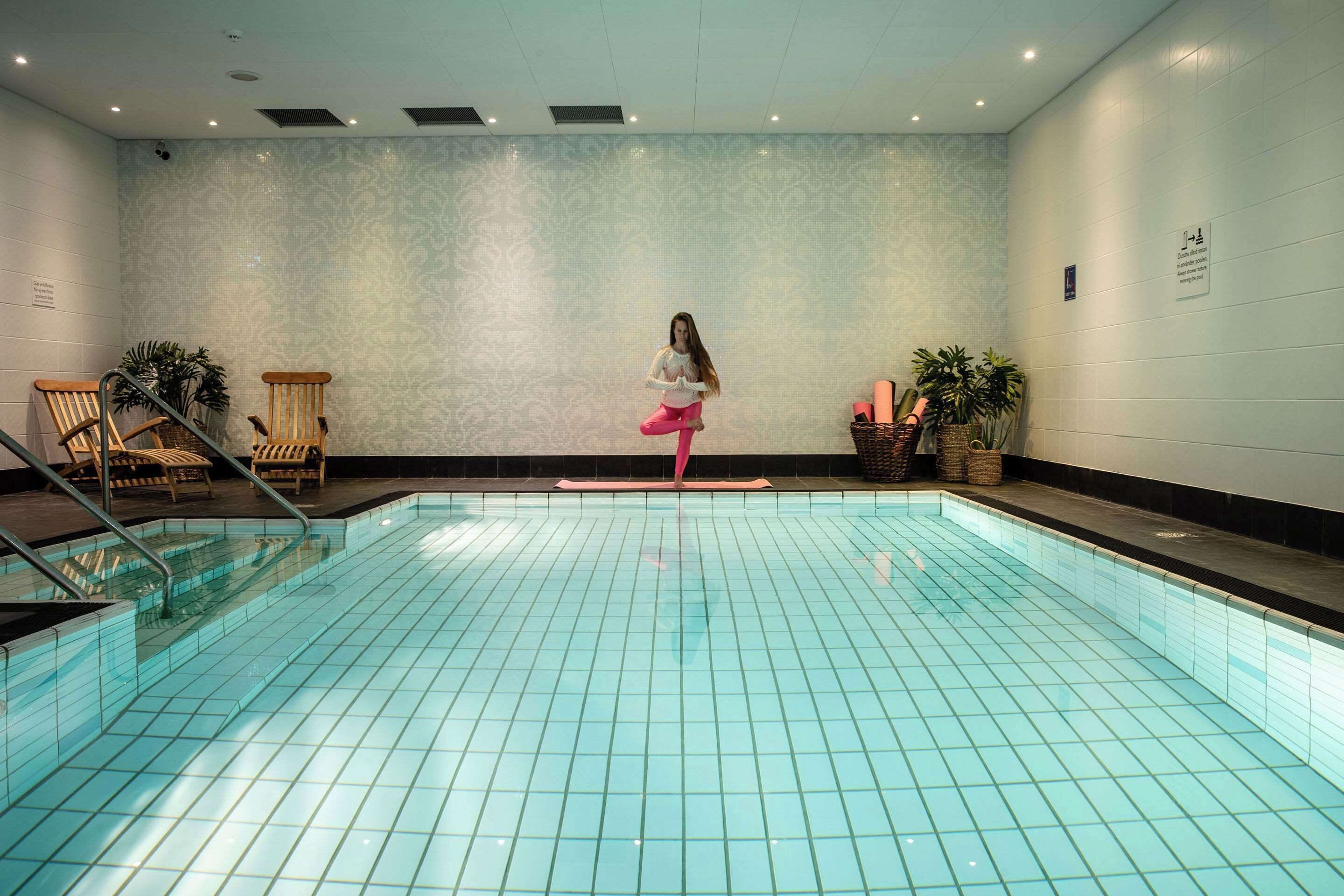 Una piscina con piso de azulejos y una persona de pie en el centro. Hay sillas y plantas en macetas alrededor de la piscina.