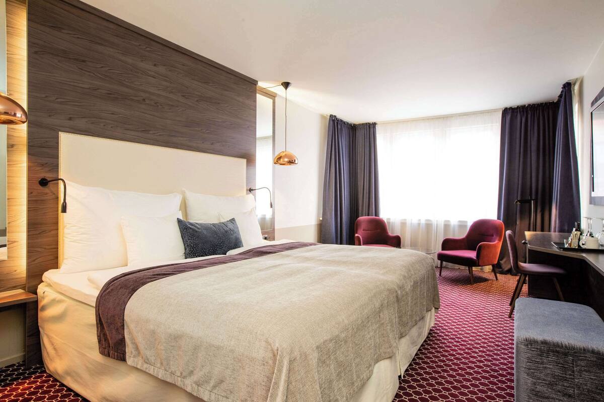 Une chambre d’hôtel avec un grand lit, un bureau, deux chaises et une fenêtre avec des rideaux.