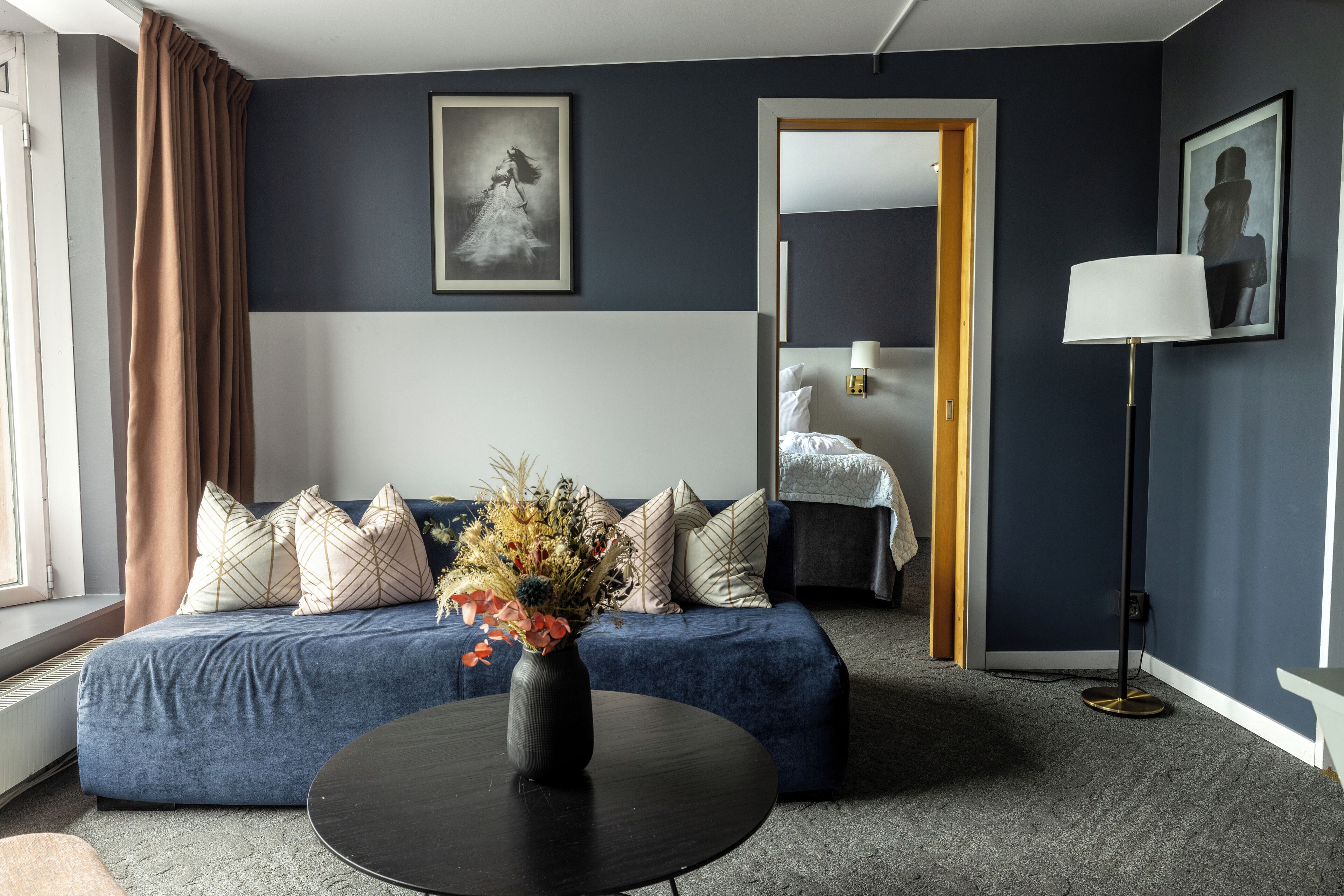 A modern hotel room with a blue sofa, a round coffee table with a vase of flowers, and a large mirror.